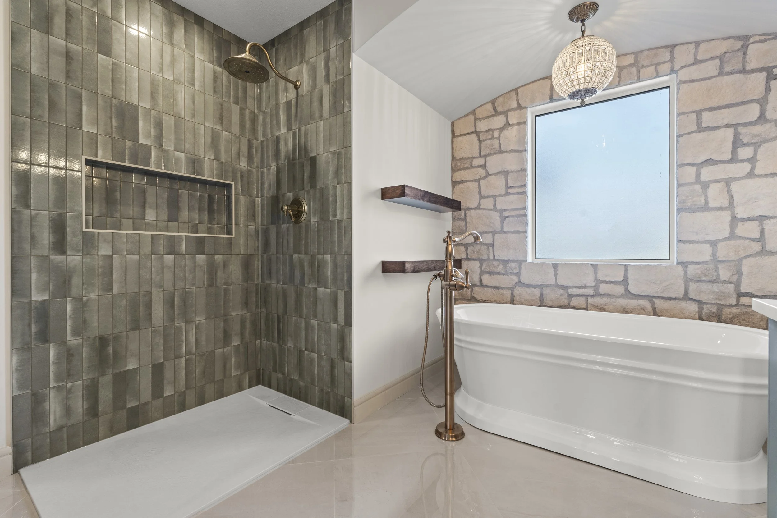 Bathroom with walk-in shower featuring dark gray tiles, built-in shelf, brass showerhead, and a freestanding bathtub near a stone accent wall and a large frosted window, with a decorative hanging light fixture.