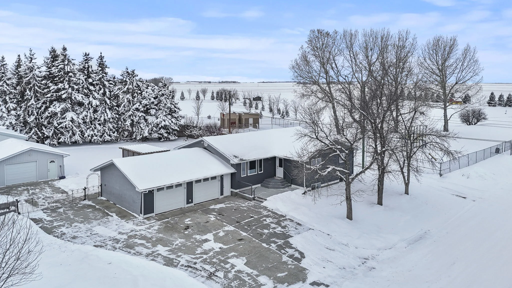 A snow-covered suburban house with a driveway and three-garage doors, next to leafless trees and a fenced yard, with snow on the roof and surrounding landscape, under a cloudy sky.