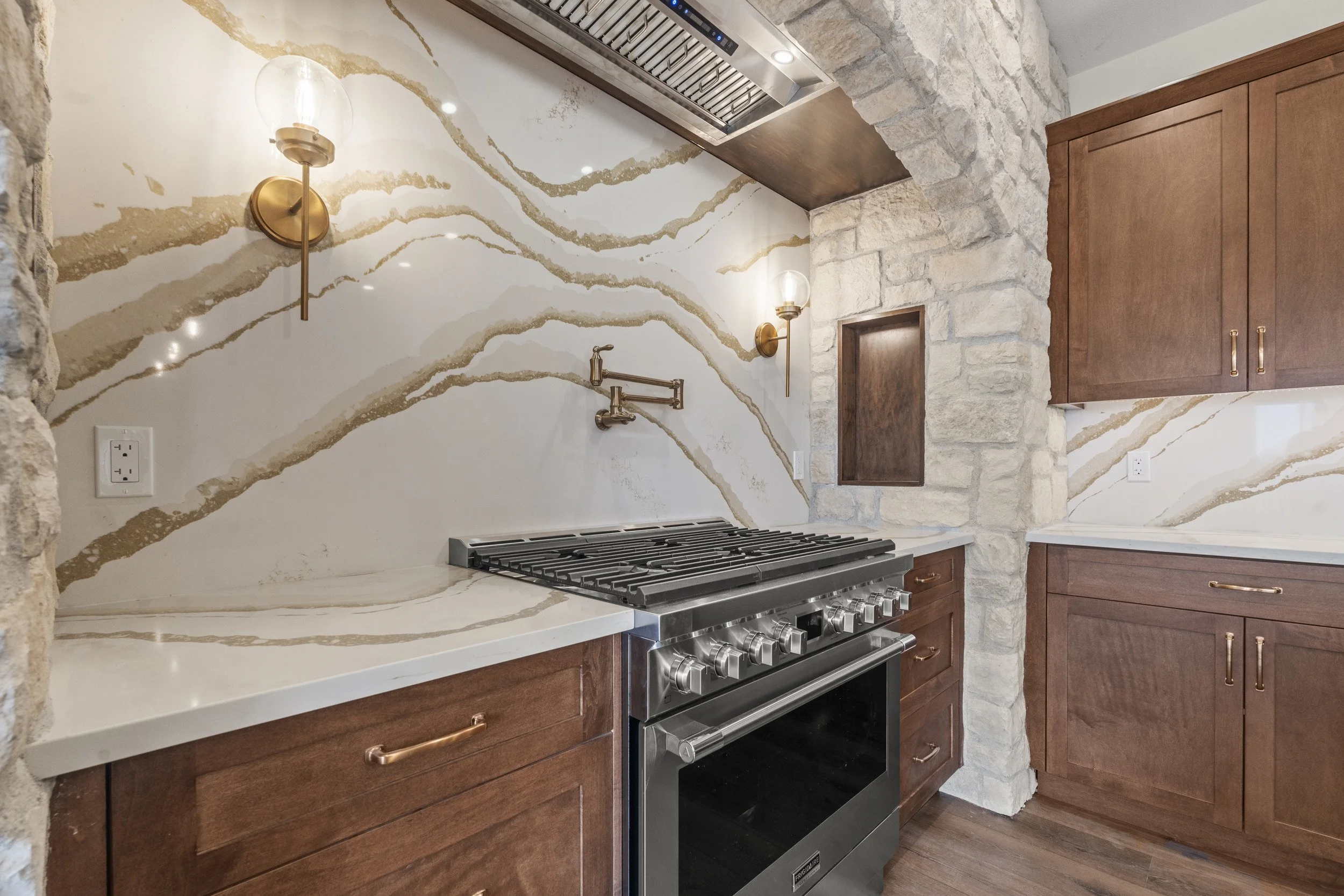 A modern kitchen with a white and gold marble backsplash, stone wall accents, and wooden cabinetry. There are brass wall sconces and a stainless steel stove.