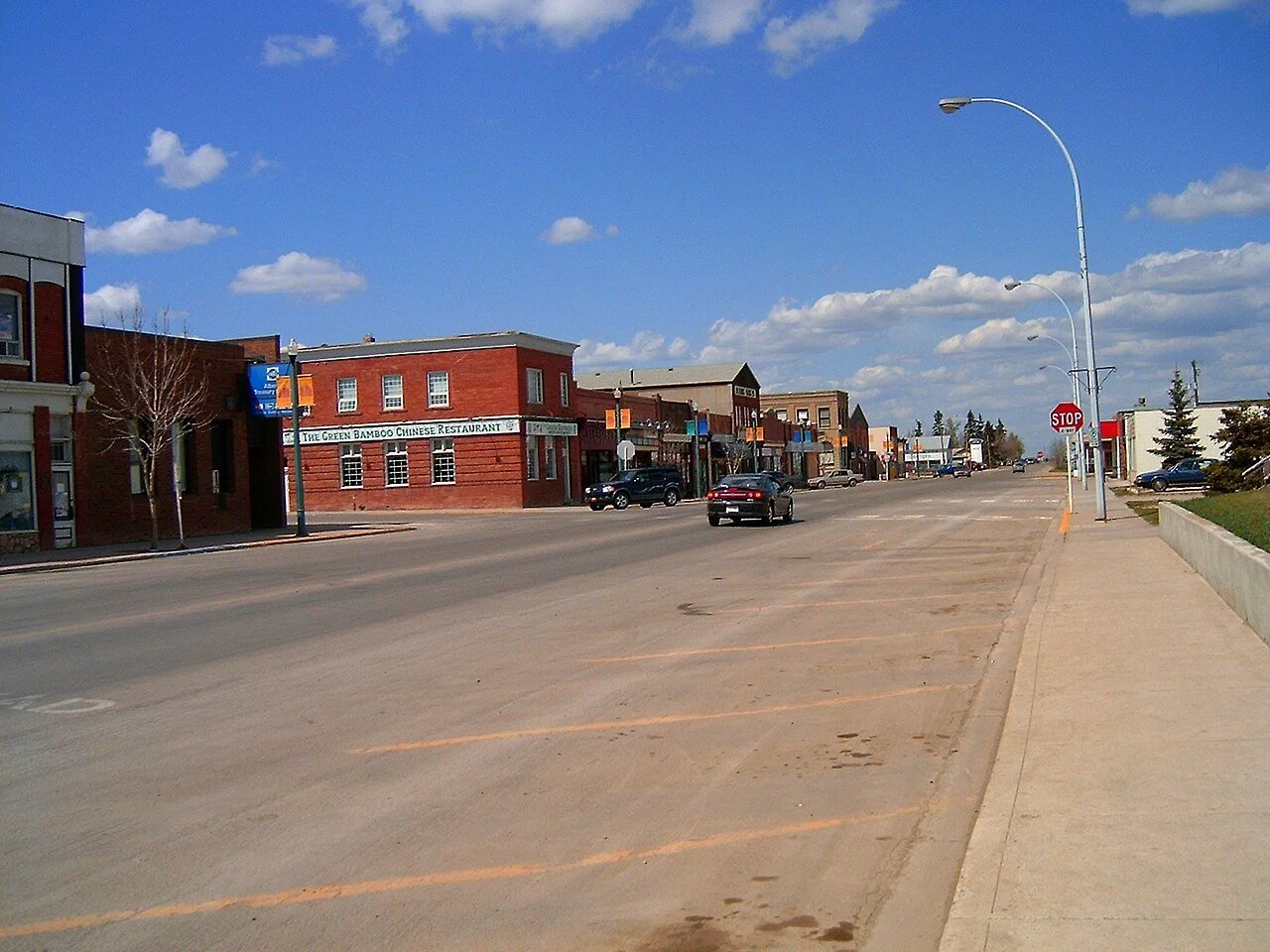 A small downtown street with shops and businesses, including a Chinese restaurant, under a blue sky with scattered clouds, with a few cars parked and driving.