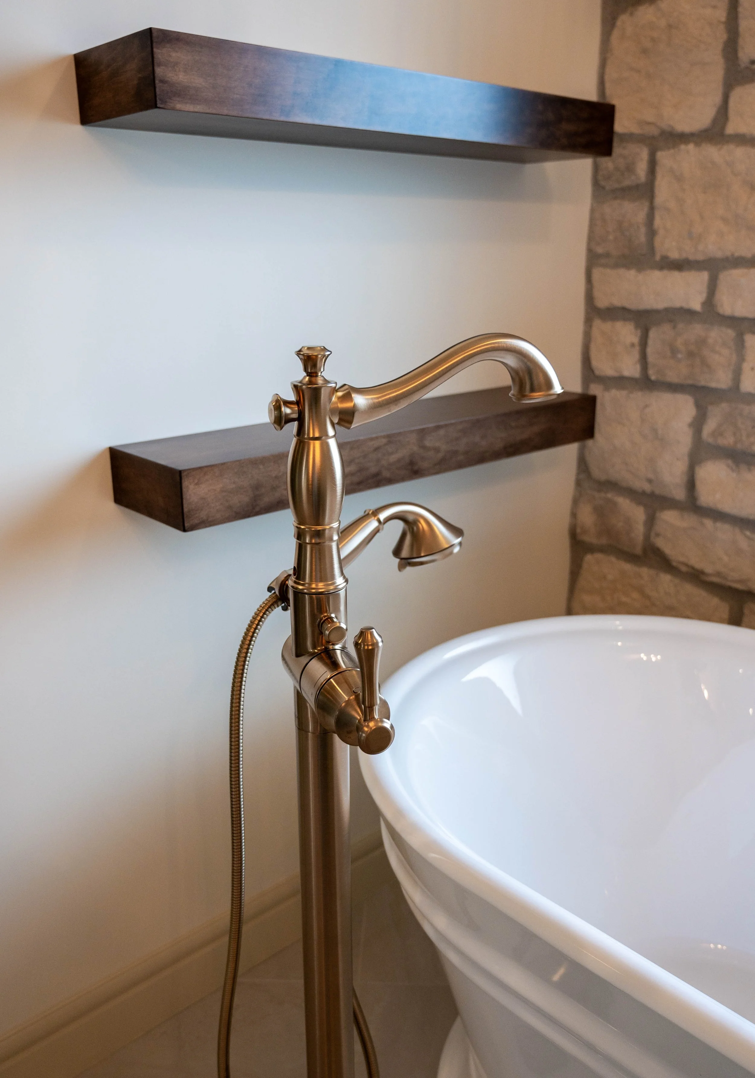 A vintage-style brass-finished bathtub faucet and handheld showerhead next to a white bathtub, with dark wood shelves and a stone wall in the background.