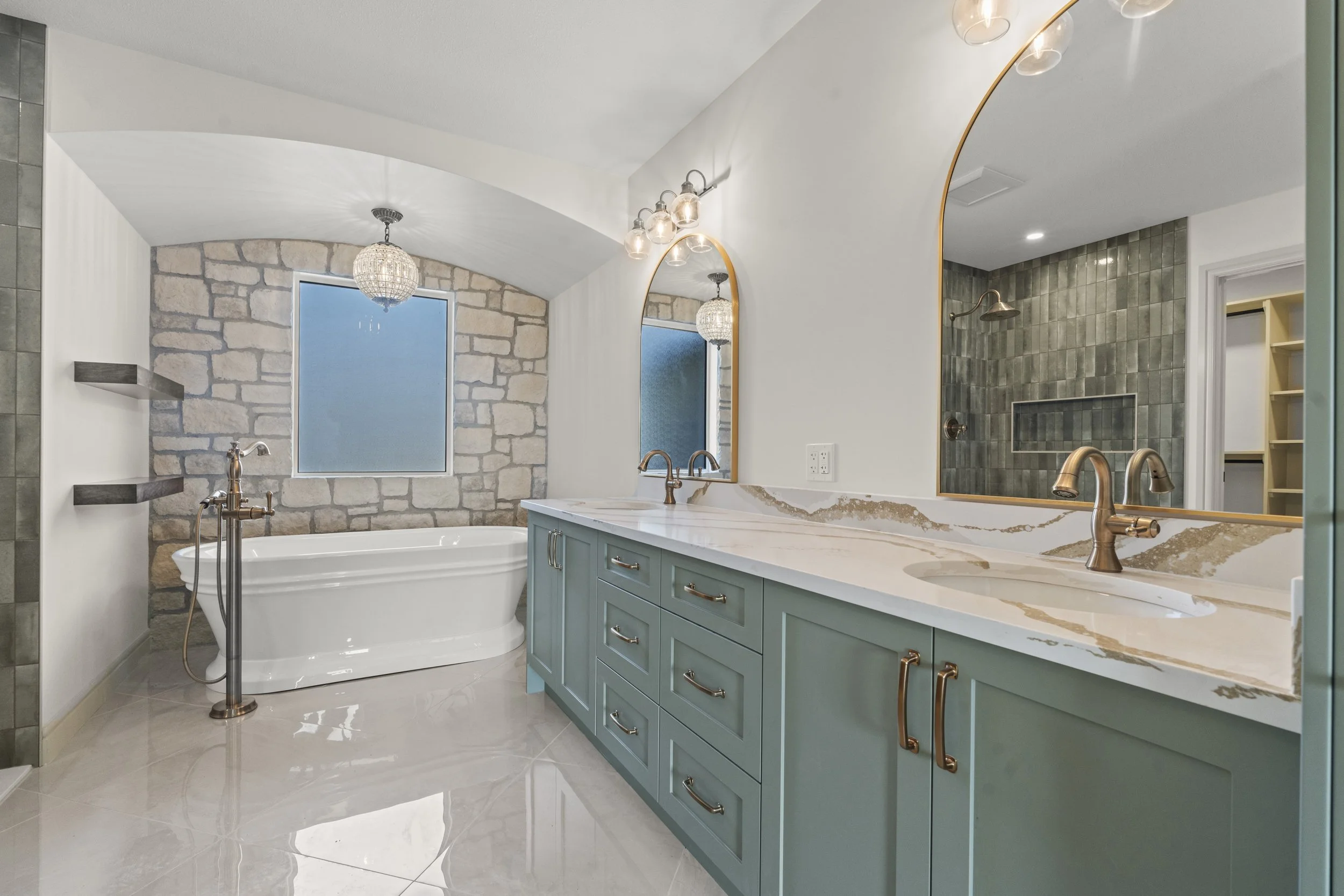 Modern bathroom with large mirror, double sink vanity with marble countertop, green cabinetry, and brass fixtures. Features a free-standing tub beneath a window with stone wall behind it, a shower with green tiles, and ambient lighting.