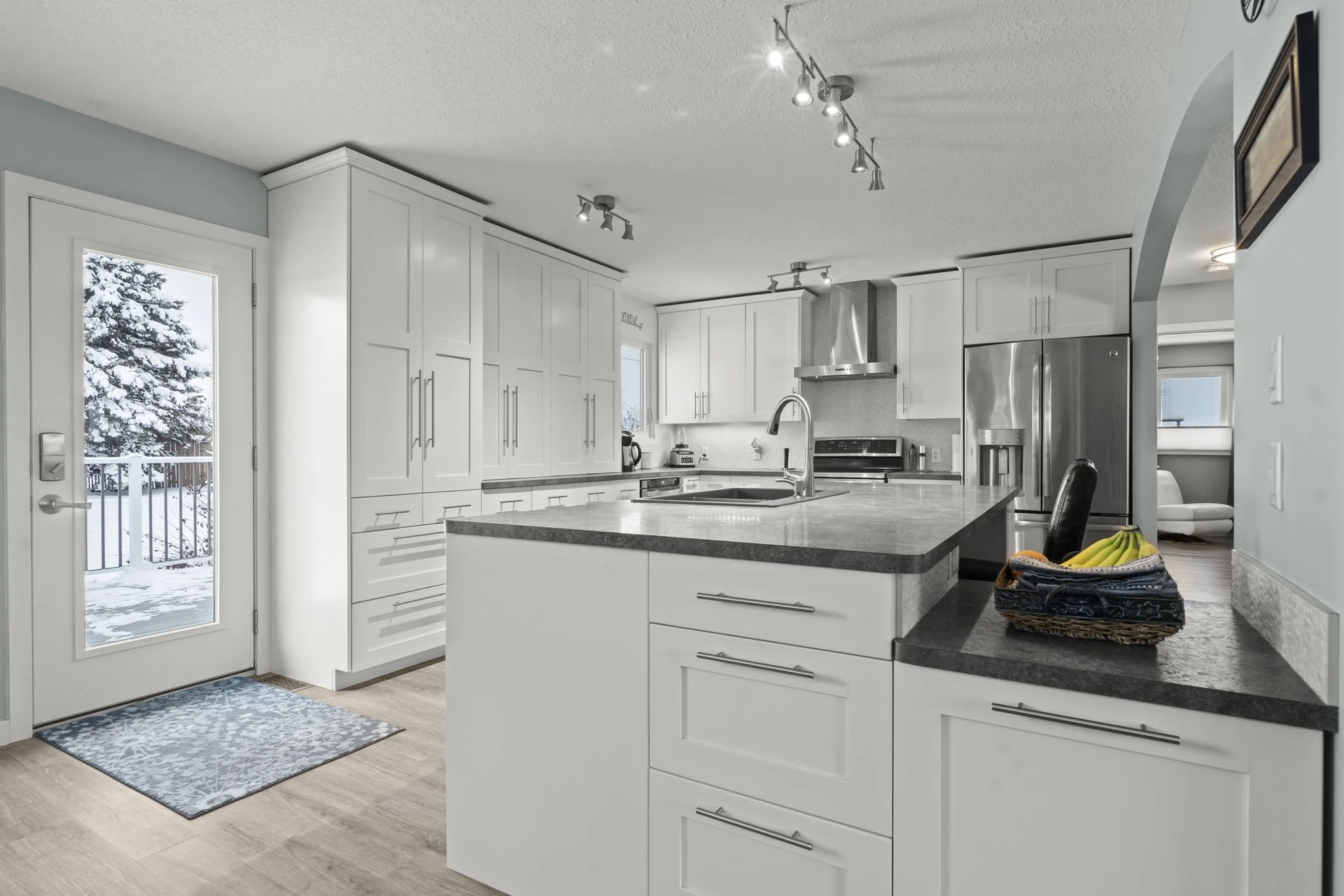 Modern kitchen with white cabinets, black countertops, stainless steel appliances, a banana basket, and a door leading outside to a snowy backyard.