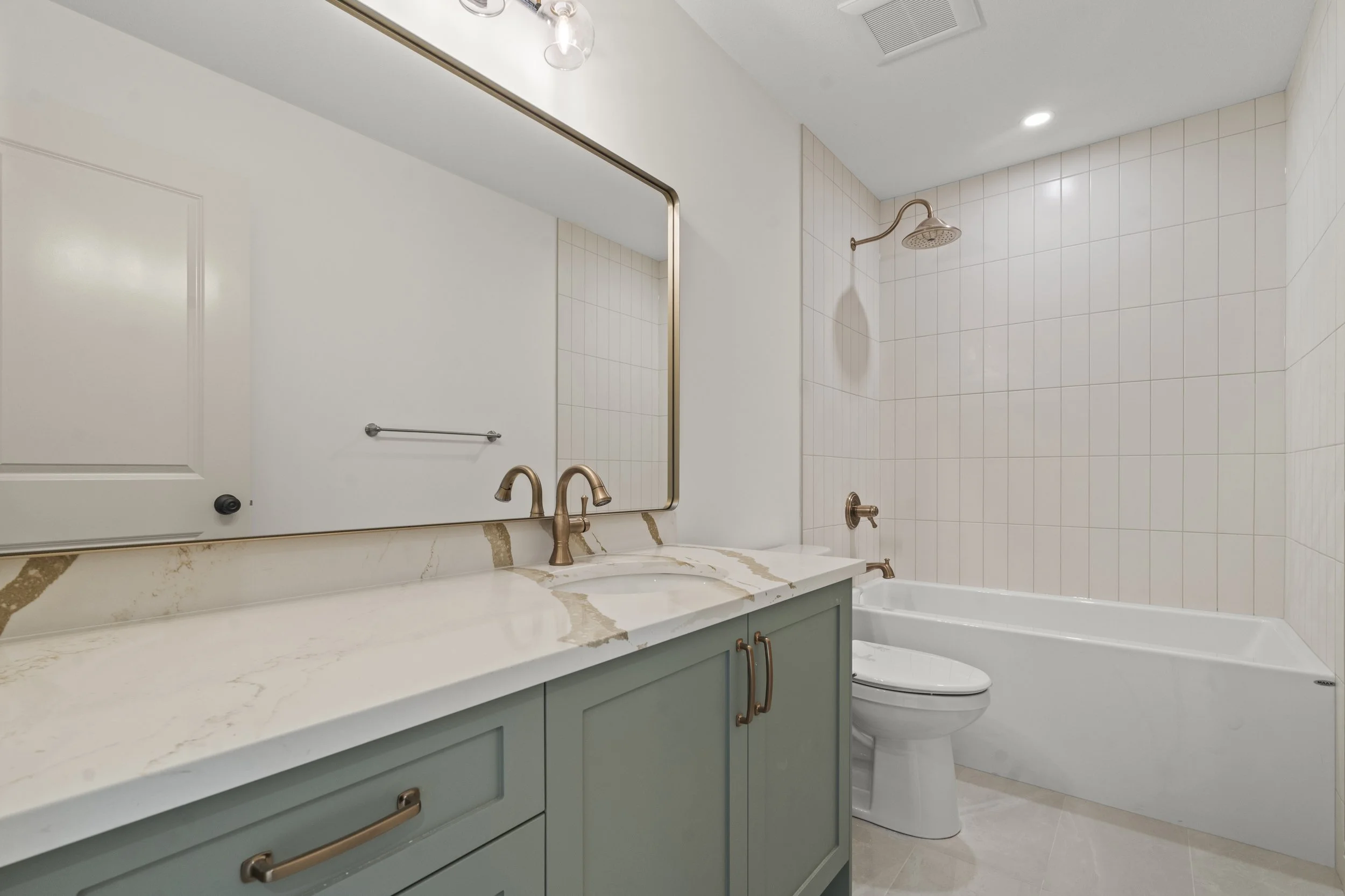 Modern bathroom with a large mirror, a marble countertop vanity with gold handles, a white toilet, and a bathtub with a showerhead in a tiled wall.