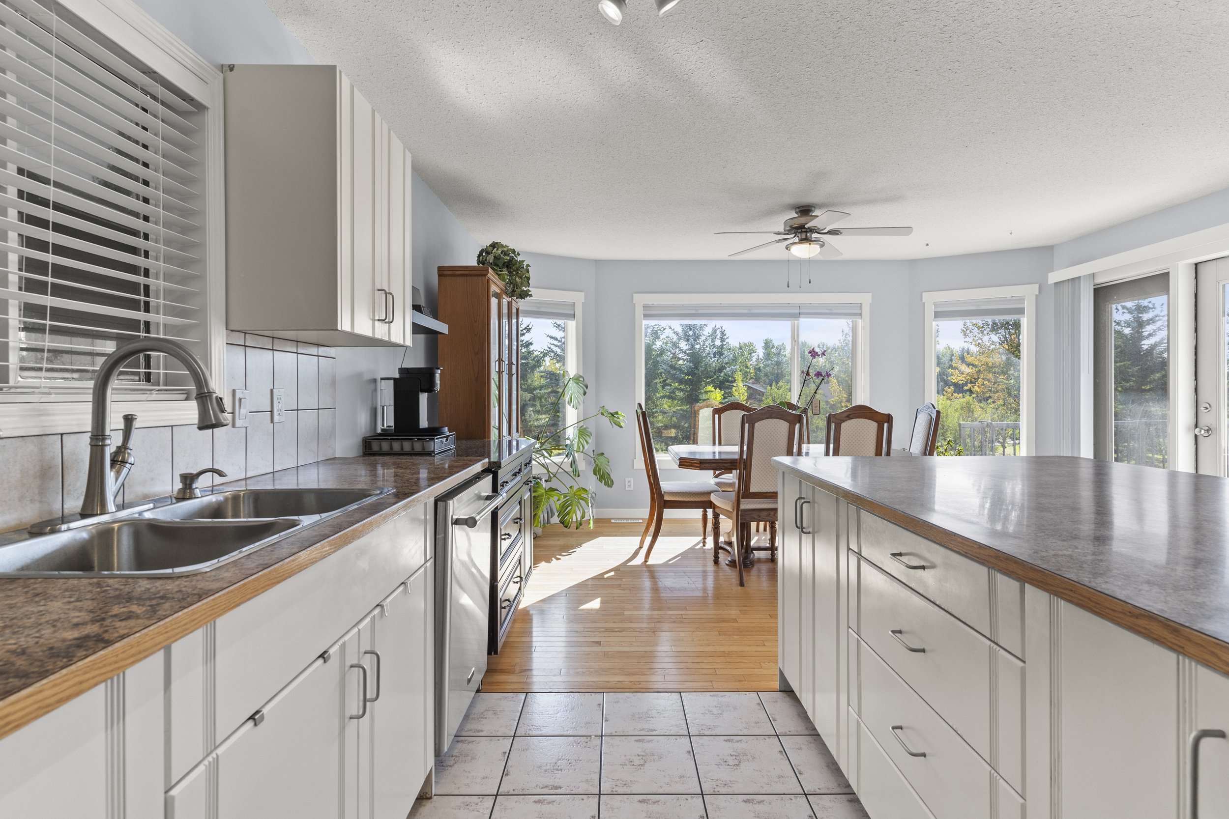 Kitchen with white cabinets, granite countertops, stainless steel sink, and a dining area with wooden table and chairs in front of large windows showing outdoor trees.