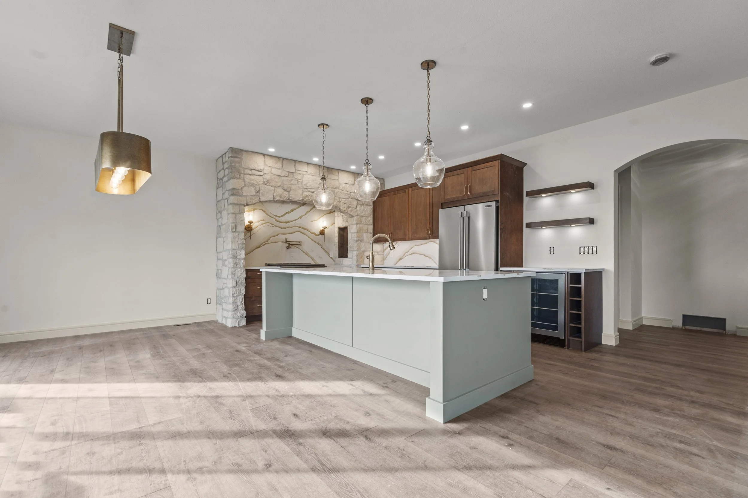 Open kitchen with a green island, wooden cabinets, stone accent wall, and hanging pendant lights.
