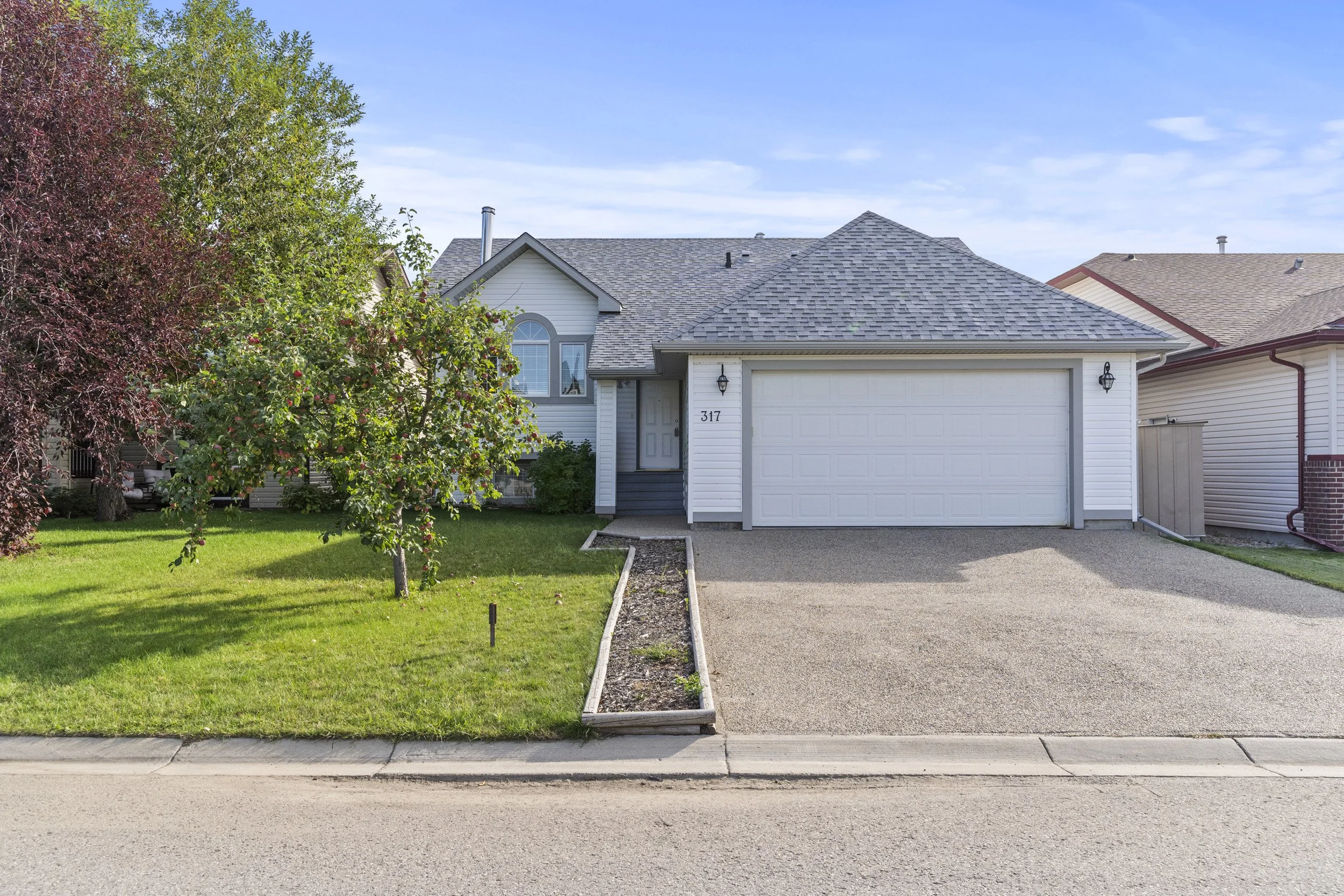 A modern suburban house with a gray shingled roof, white siding, a two-car garage, and a small front yard with grass and a tree.