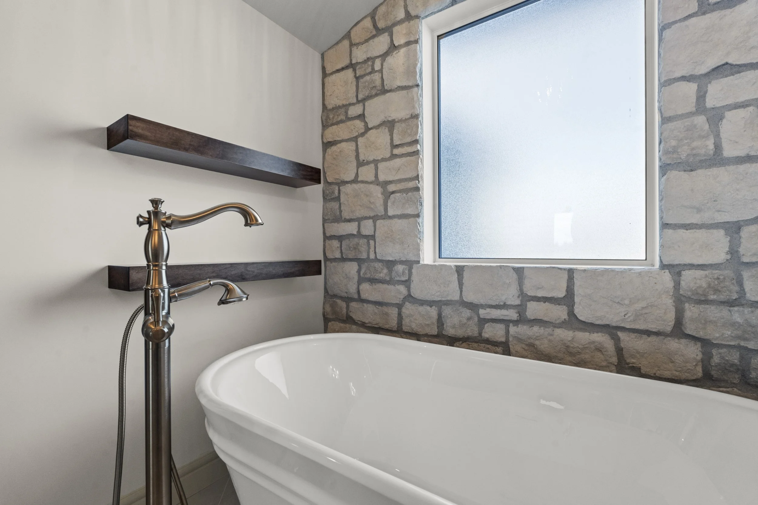 Bathroom with a white bathtub, stone wall, frosted window, and dark wood shelves.