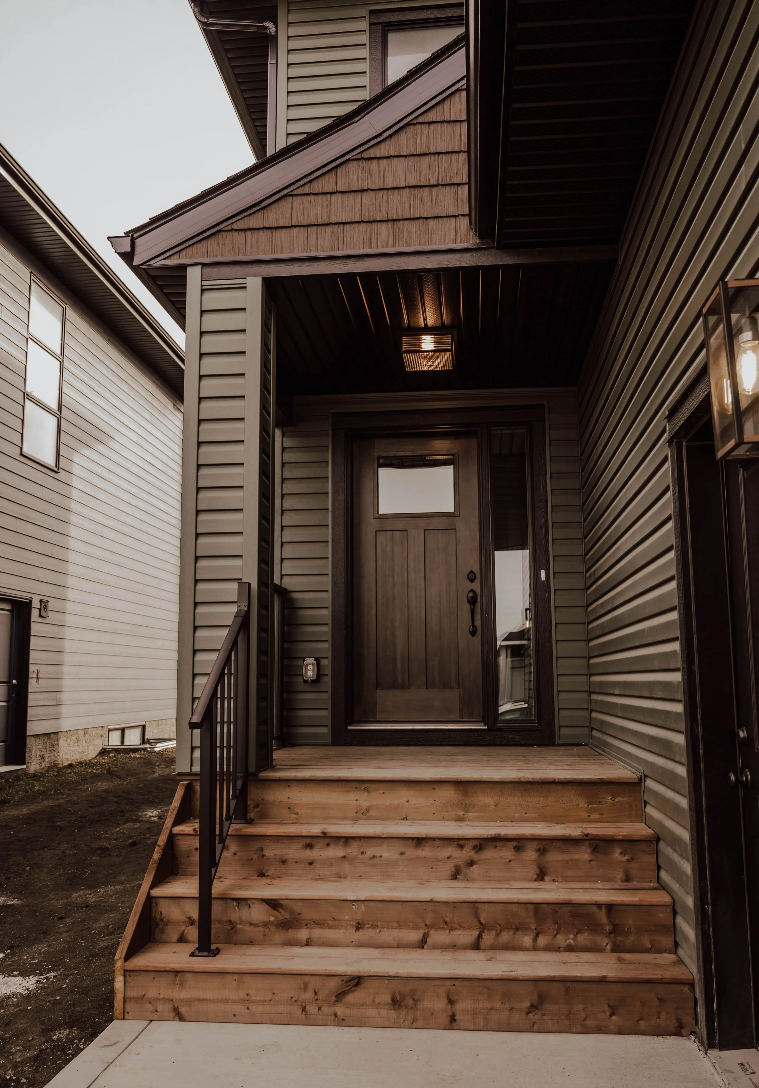 Front porch of a house with wooden stairs, black railing, and a dark brown door, with exterior siding and a light fixture above.