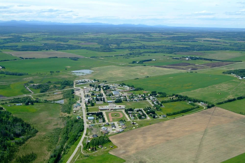 Aerial view of a small town surrounded by green fields and farmland with a large baseball field and nearby roads.