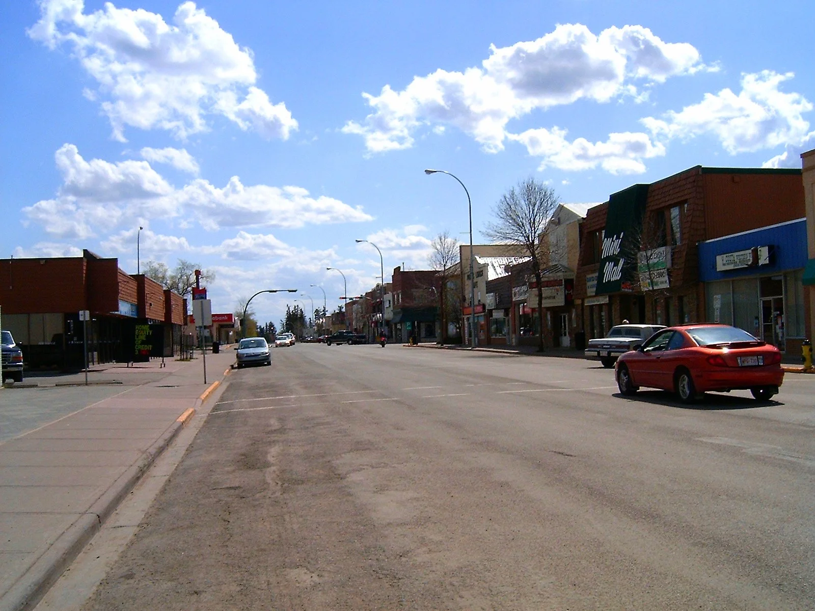 A small town street with shops and parked cars on a sunny day with a partly cloudy sky.