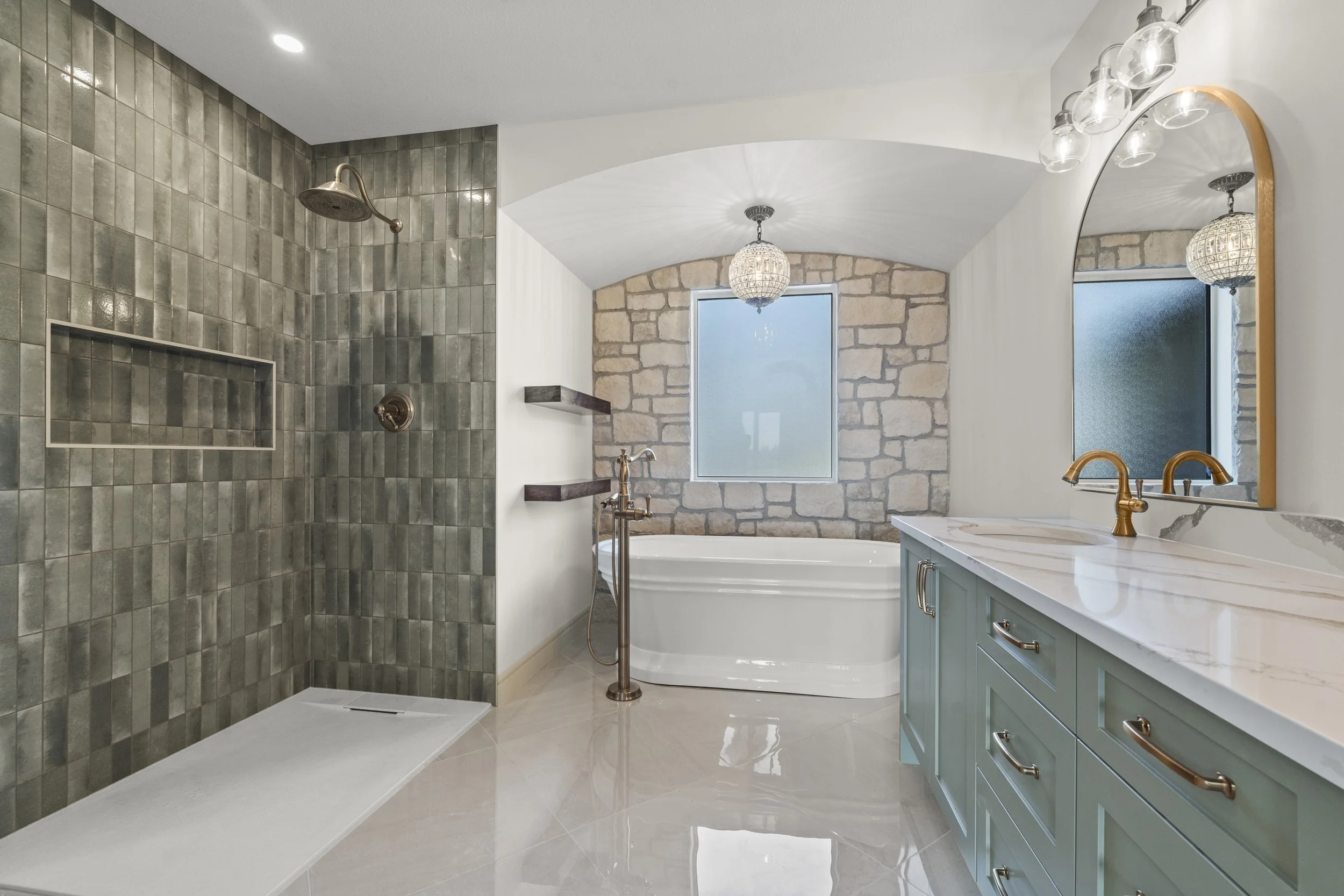 A spacious bathroom featuring a stone-tiled wall with a shower area, a freestanding bathtub under a window, a vanity with a marble countertop, gold fixtures, and decorative lighting