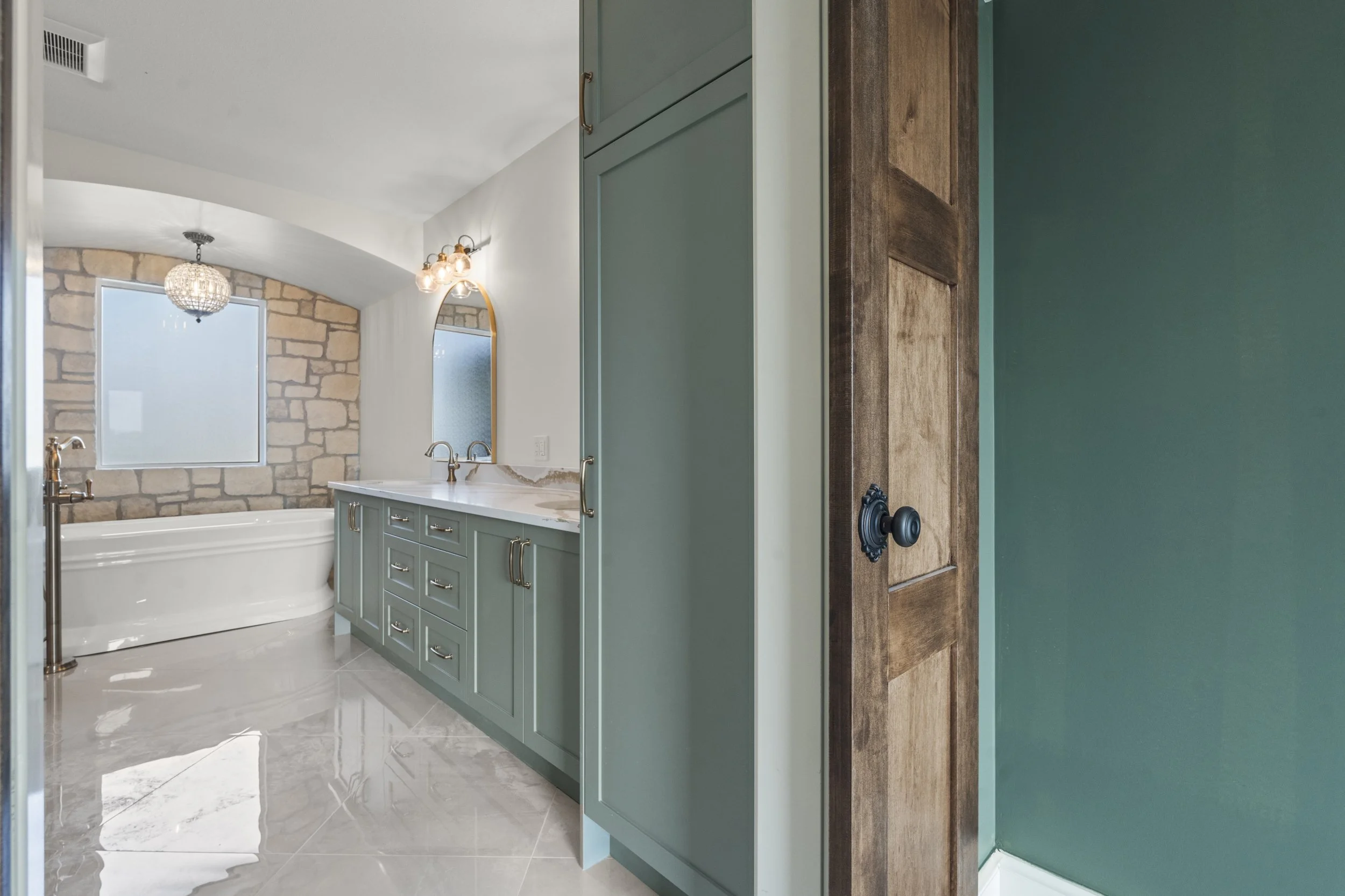Bathroom with a green vanity, marble countertop, oval mirror, sconce lighting, bathtub, frosted window, stone wall, tiled floor, and a green wall with a wooden door.