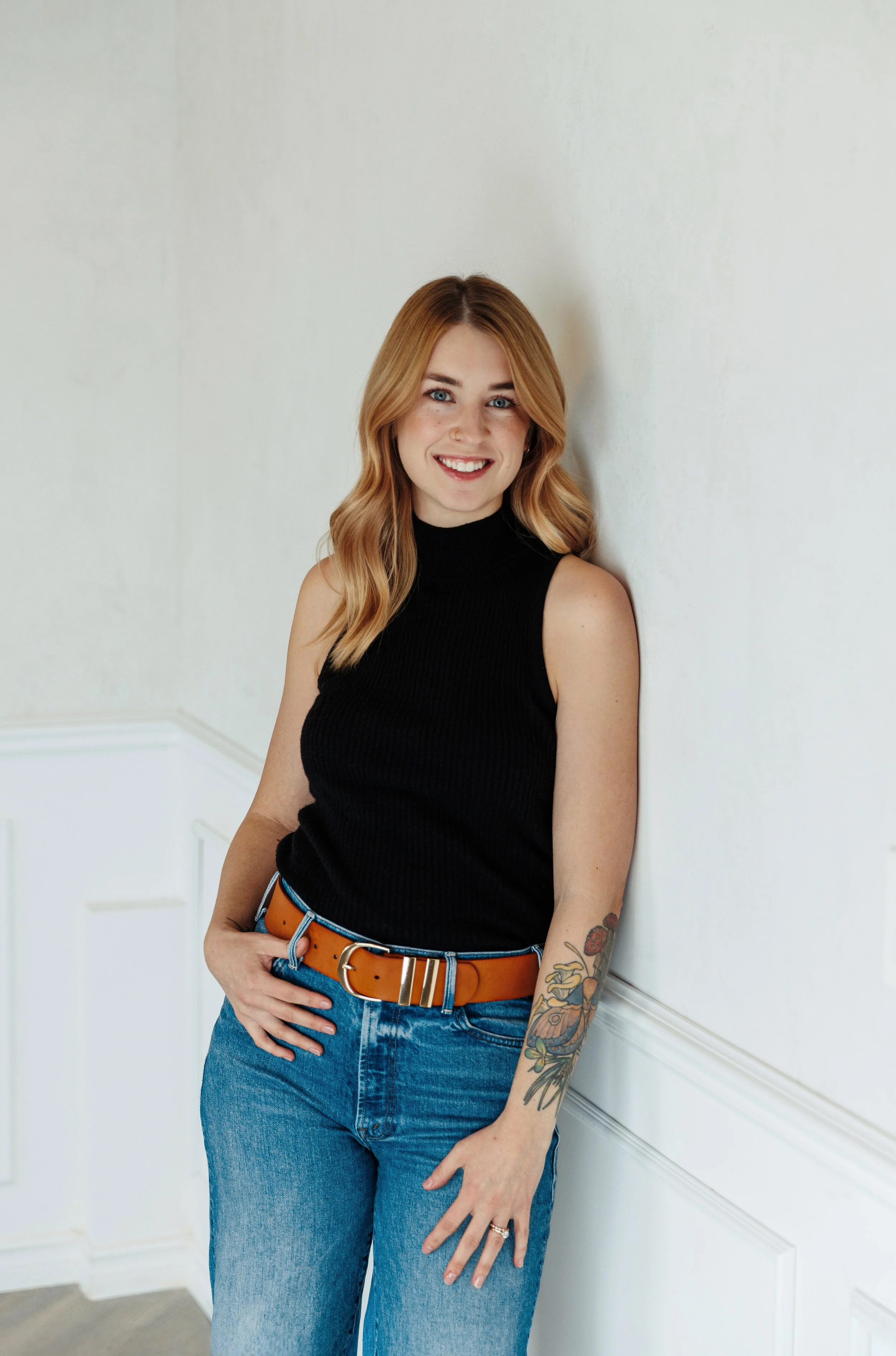 A smiling woman with blonde hair wearing a black sleeveless turtleneck top and blue jeans, standing against a light-colored wall with decorative molding.