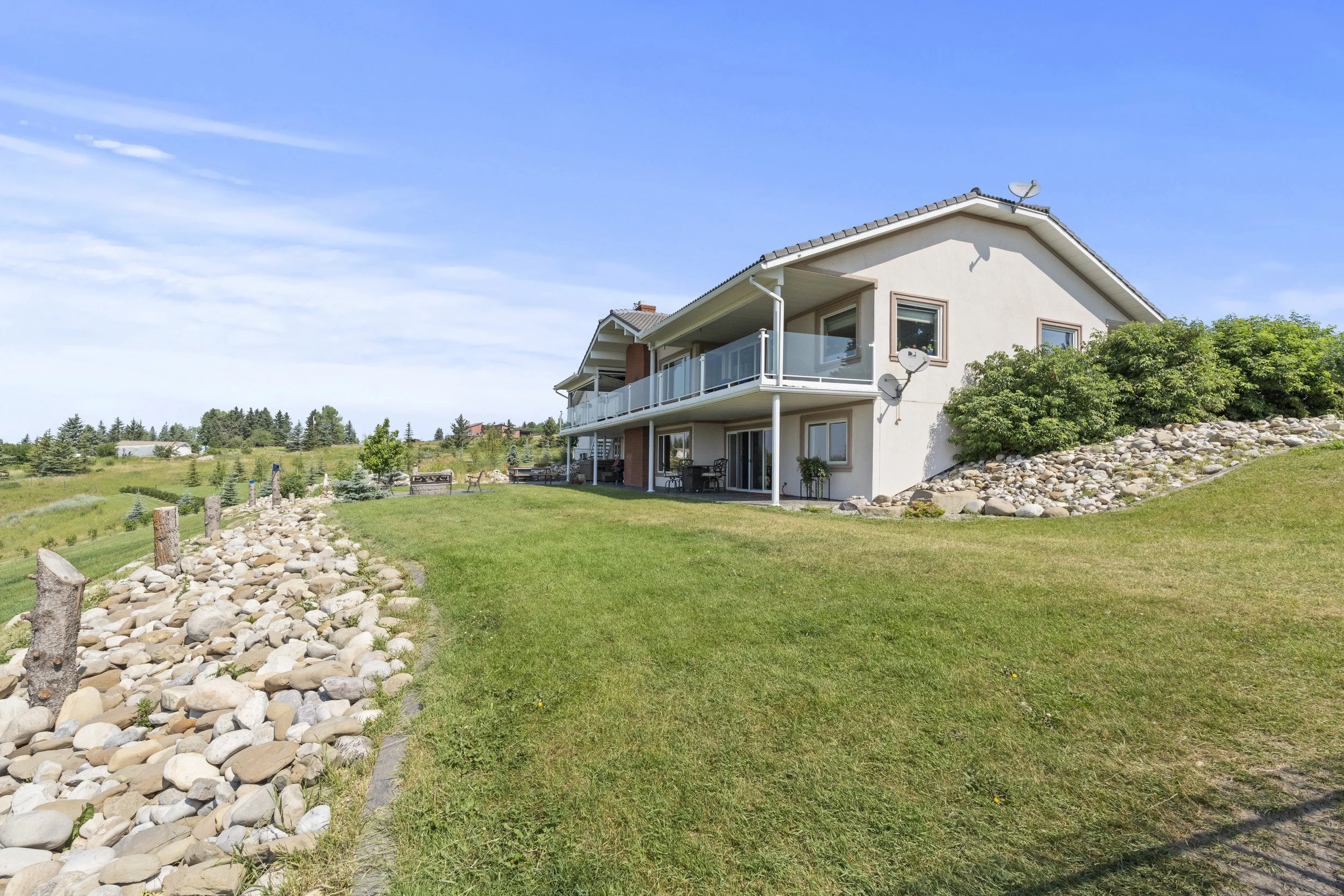 A two-story house with a balcony on the upper floor, satellite dishes on the exterior, surrounded by a green lawn, rocks, and distant trees under a blue sky.