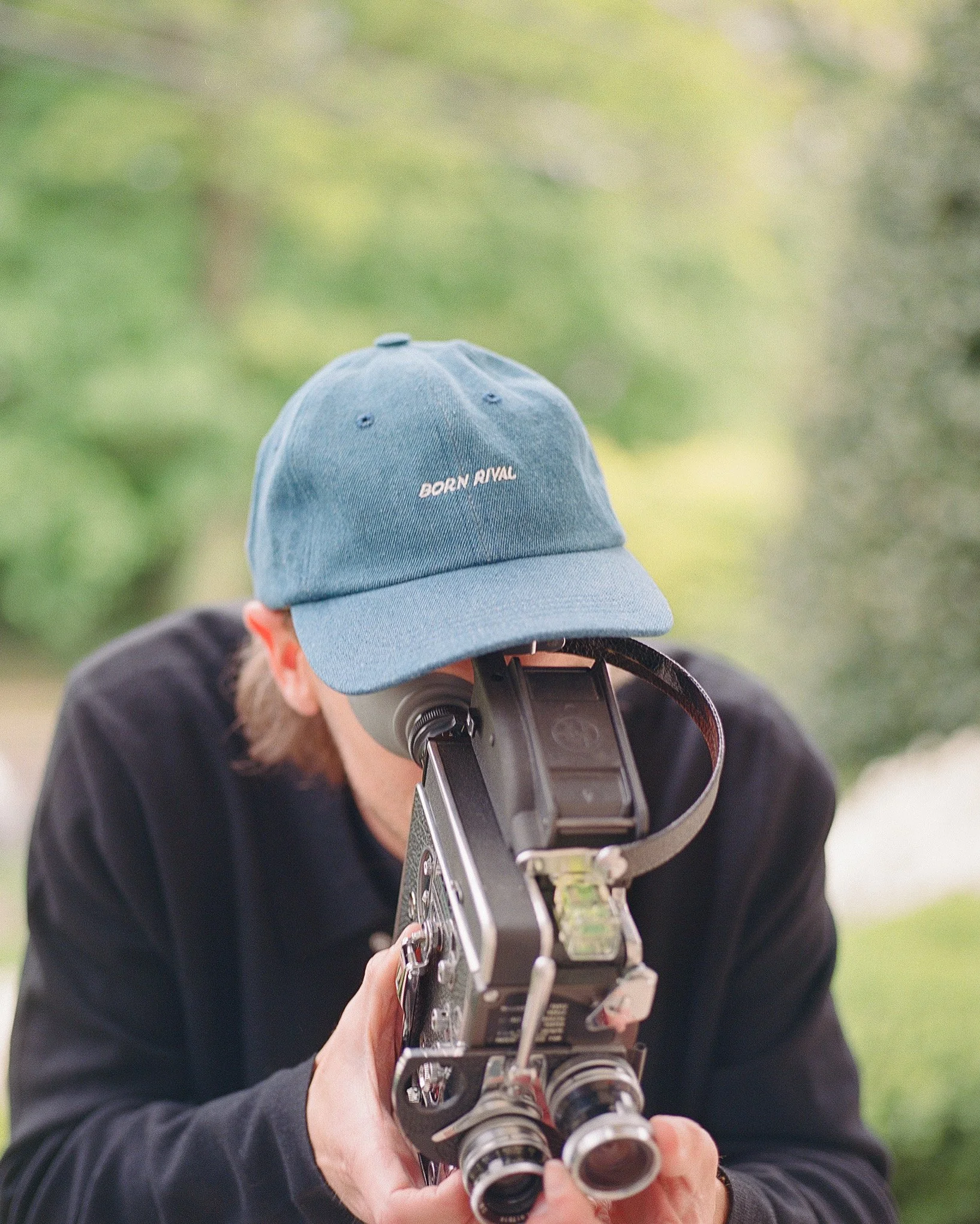 Person wearing a blue cap with 'Born Rival' text, holding a vintage film camera outdoors.