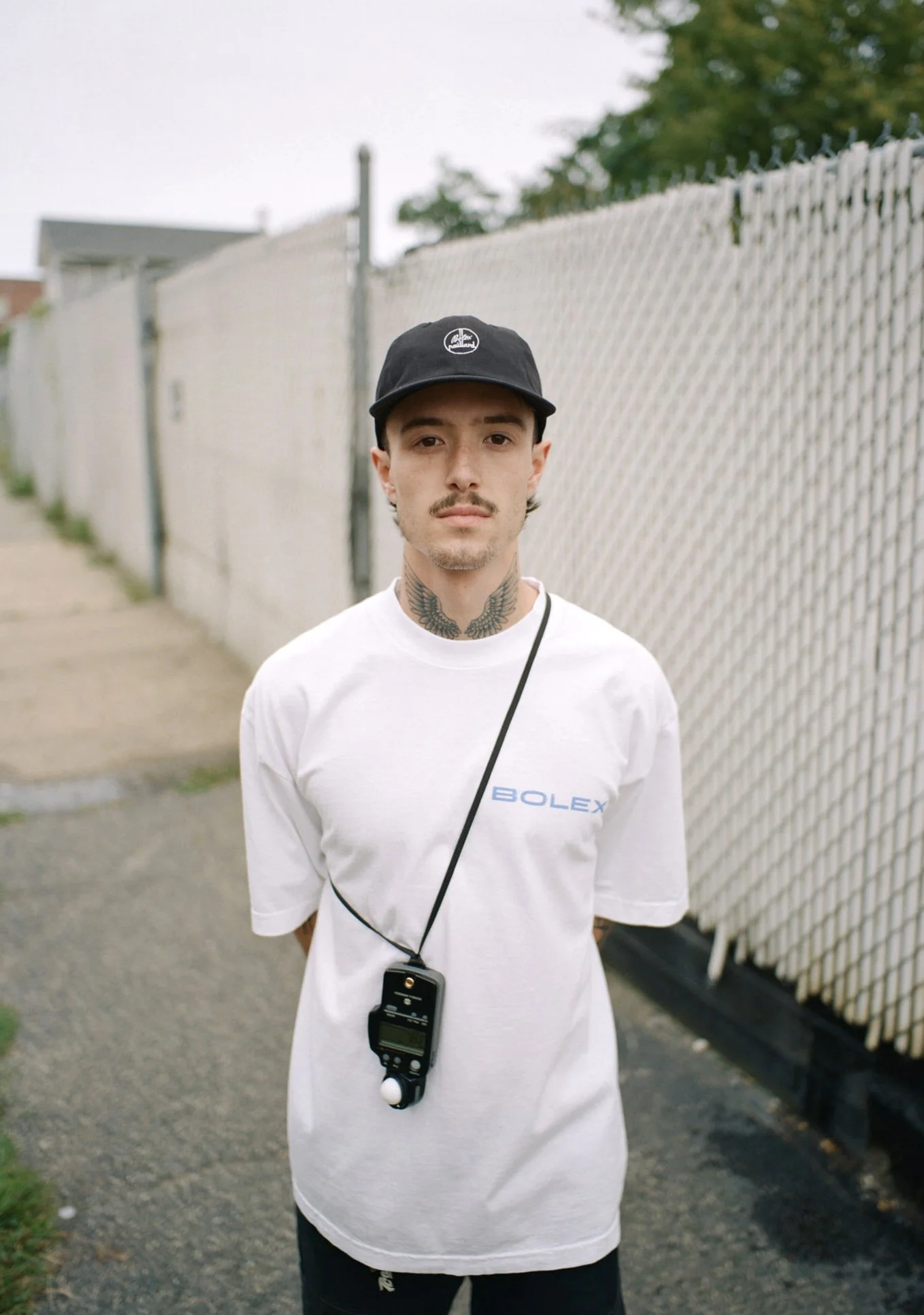 A young man with tattoos on his neck, wearing a black cap, white T-shirt with 'BOLEX' logo, and a stopwatch hanging around his neck, standing outdoors beside a chain-link fence.