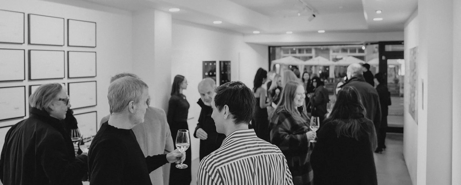 Menschen bei einer Kunstausstellung, einige trinken Wein, in einem weißen Raum, schwarz-weiß Bild.