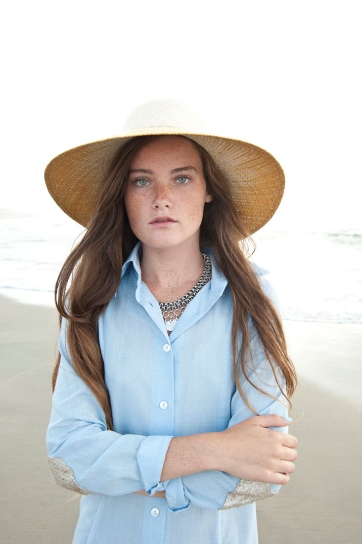 Woman in a wide-brim straw hat and a blue button-down shirt, arms folded
