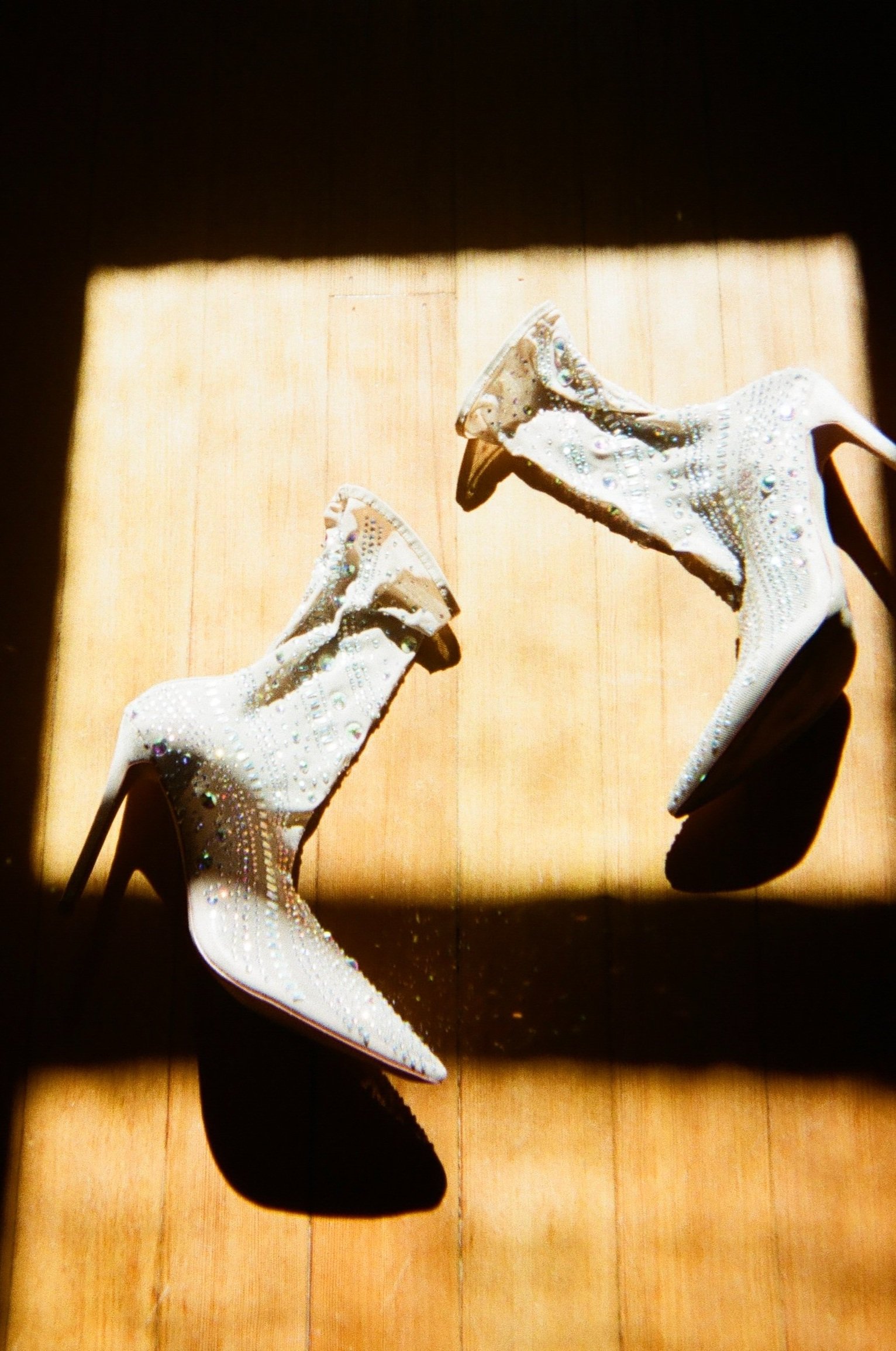Pair of white high-heeled ankle boots decorated with rhinestones and embellishments, placed on a wooden floor with sunlight creating shadows.
