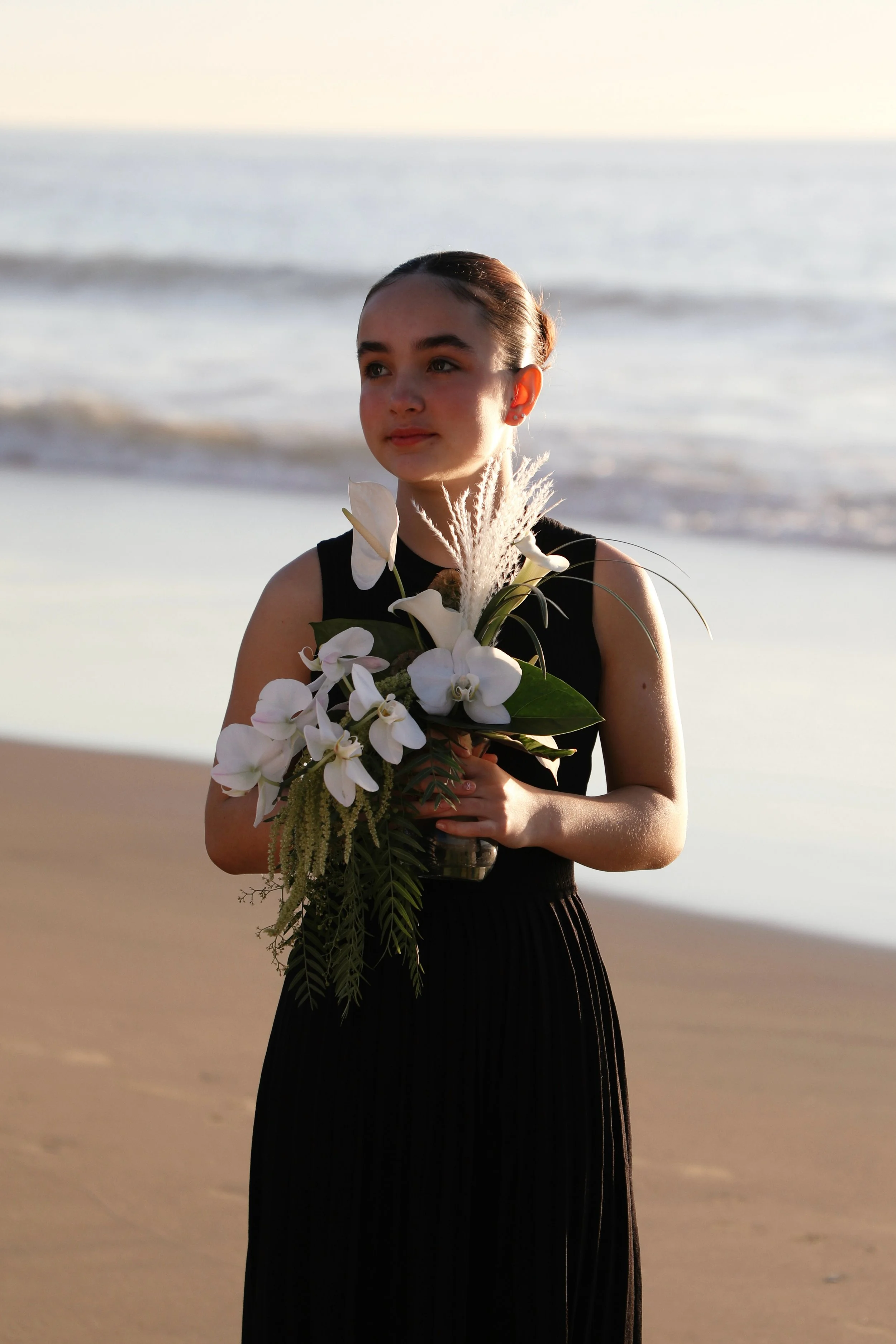 A young woman in a black dress holding a bouquet of white flowers on a beach at sunset.