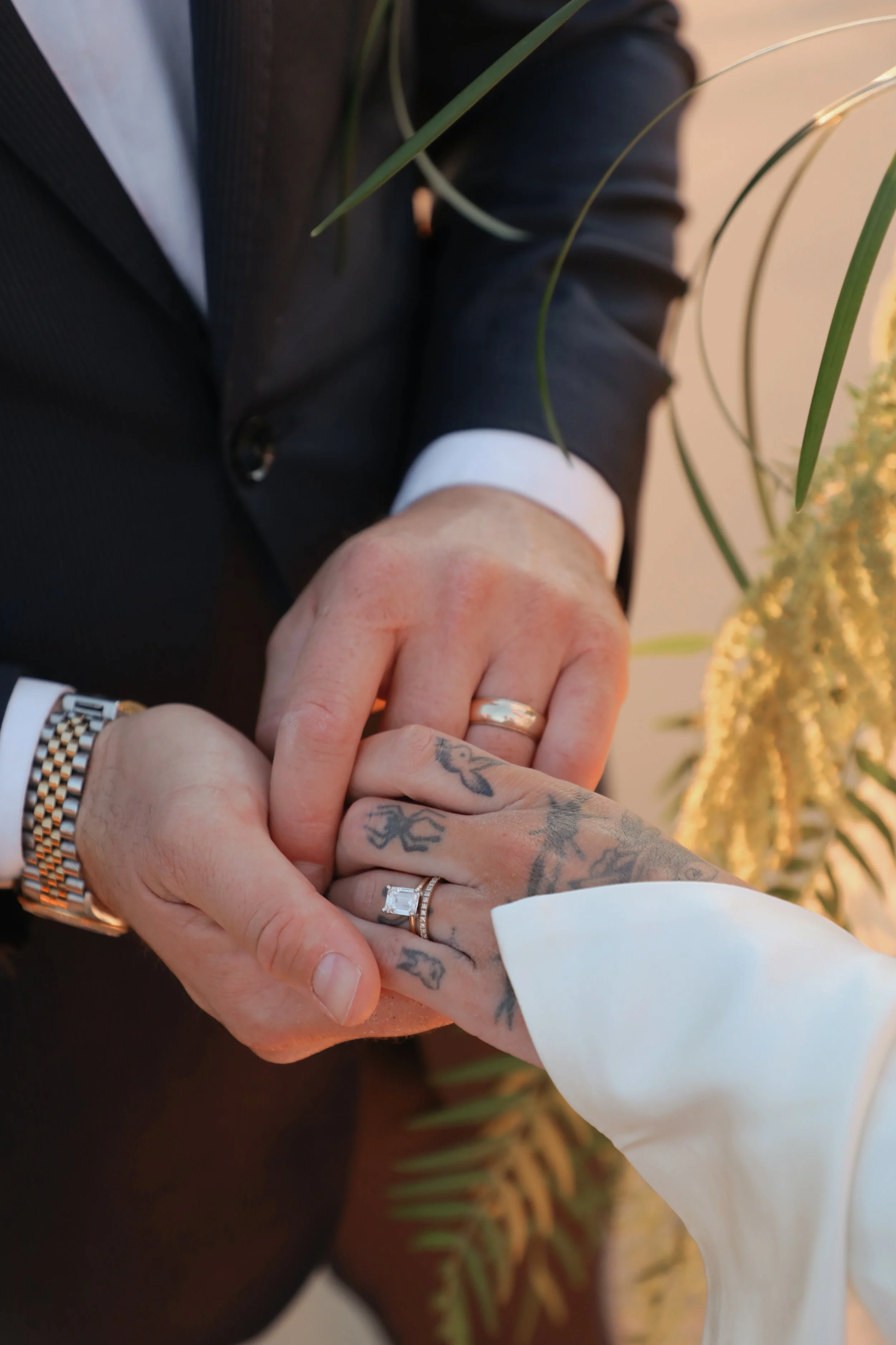 Close-up of two people holding hands, showing wedding rings and tattoos, with one person in a suit and the other in a white sleeve.