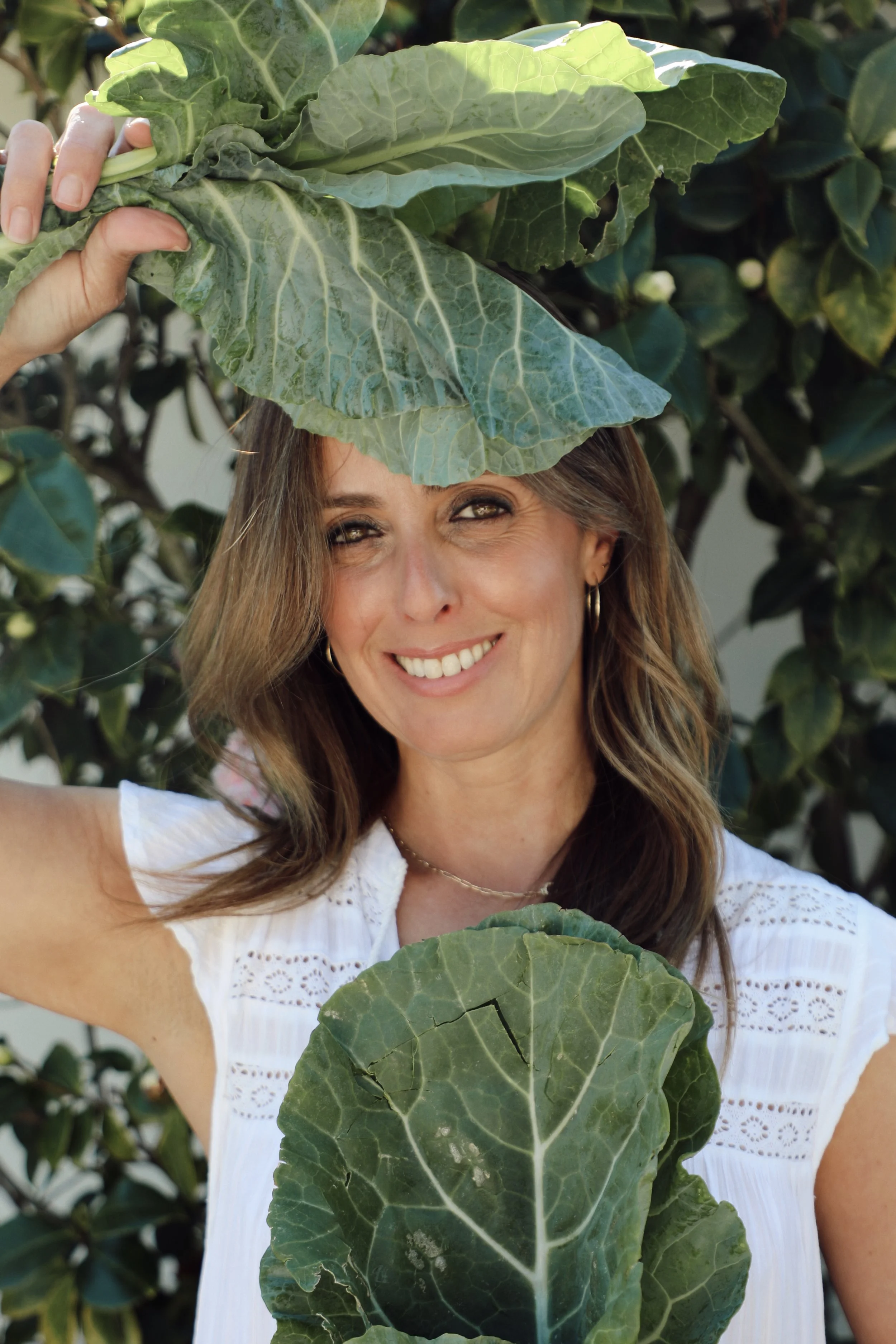 A woman smiling and holding large green leaves in front of a leafy background.