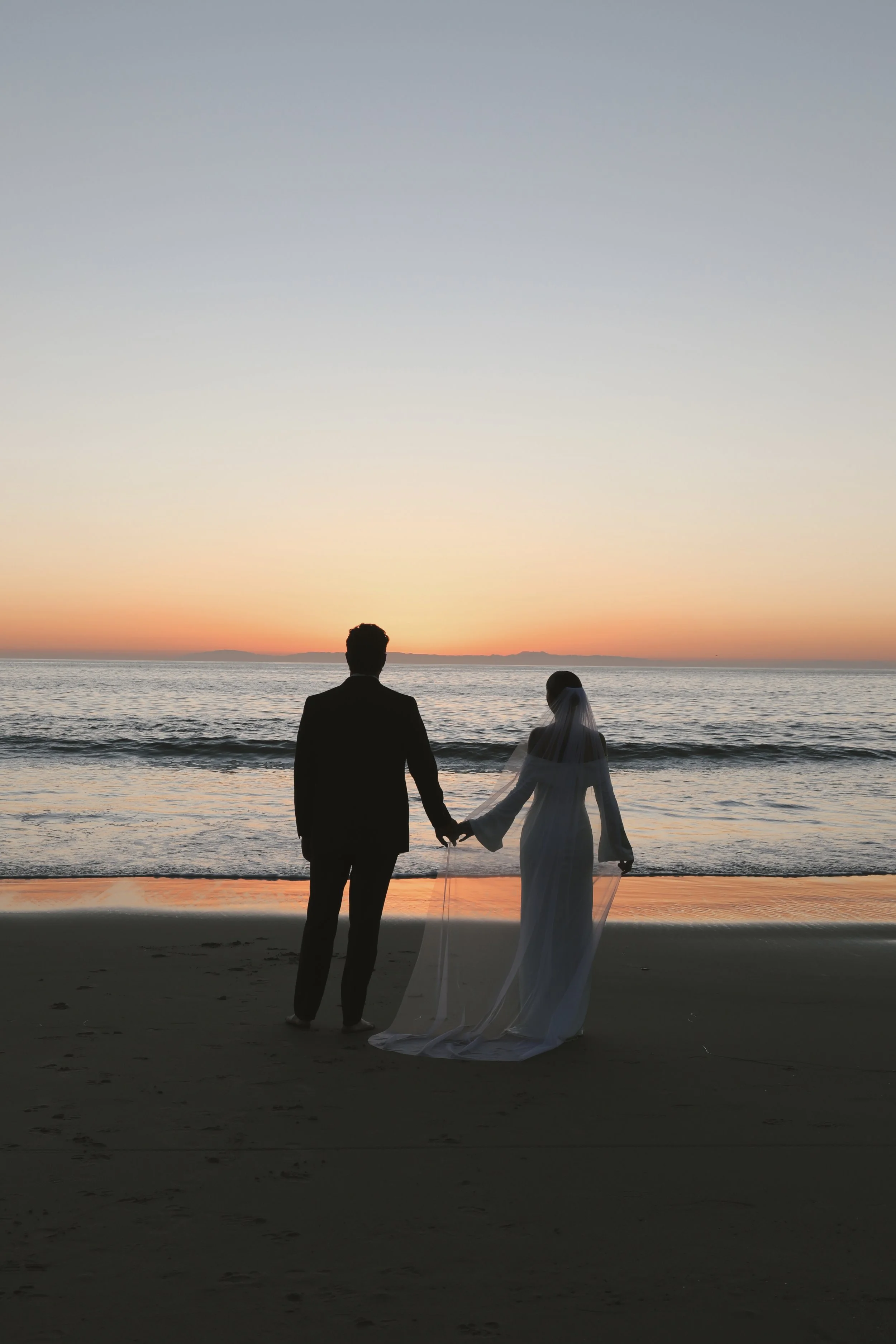 A couple dressed in wedding attire holding hands on the beach at sunset.