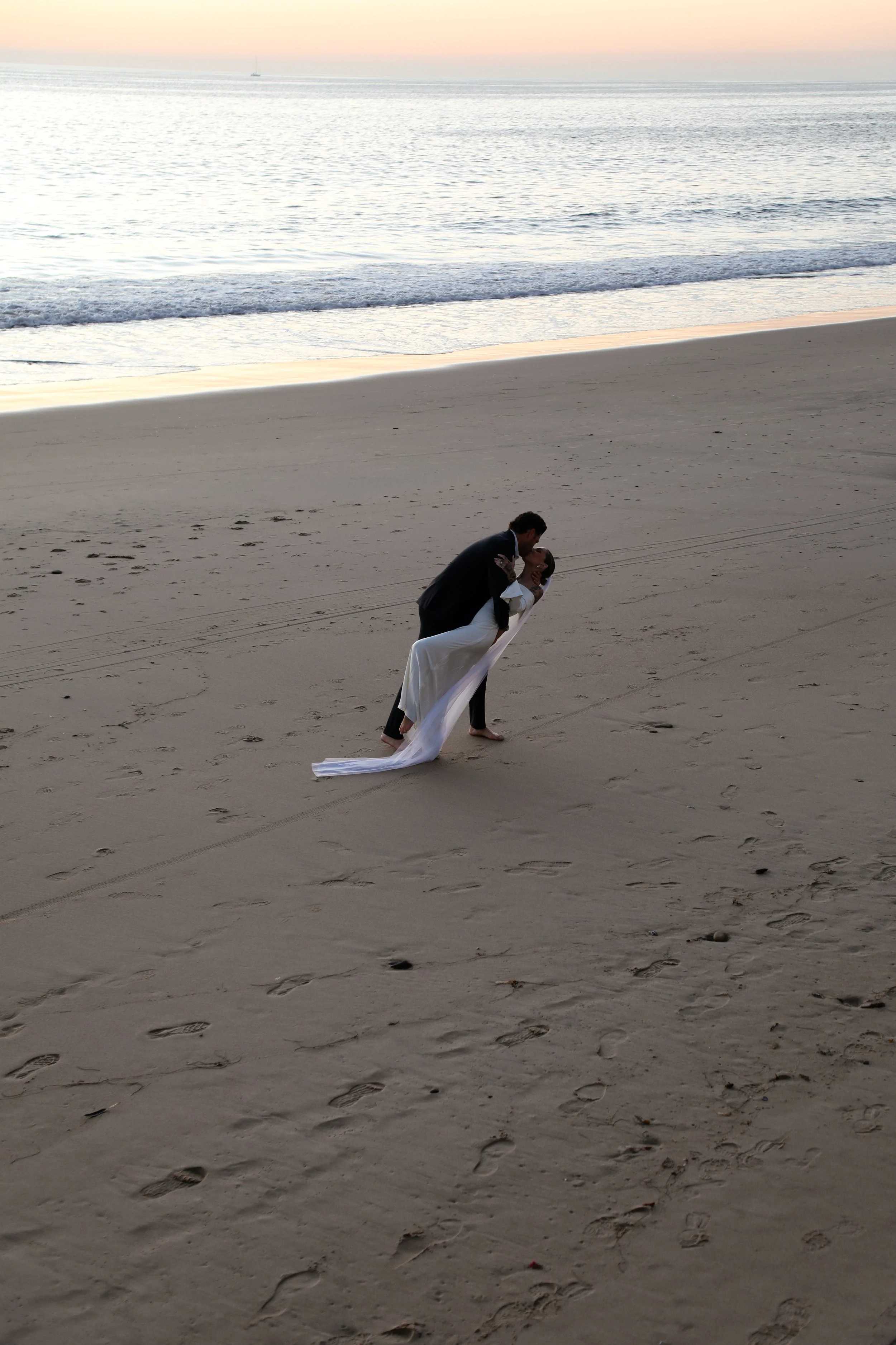 A couple in wedding attire sharing a kiss on a sandy beach at sunset