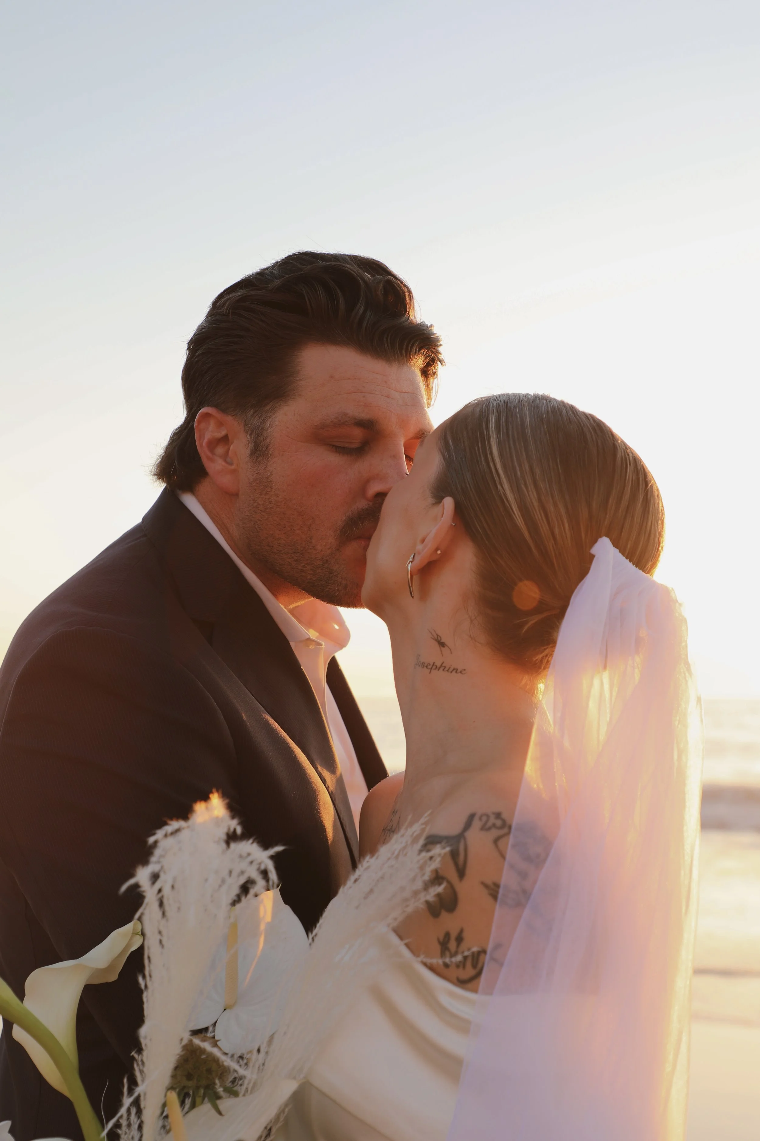 A couple in wedding attire sharing a kiss on the beach during sunset, with the woman wearing a veil and the man in a tuxedo, surrounded by white flowers and tall grass.
