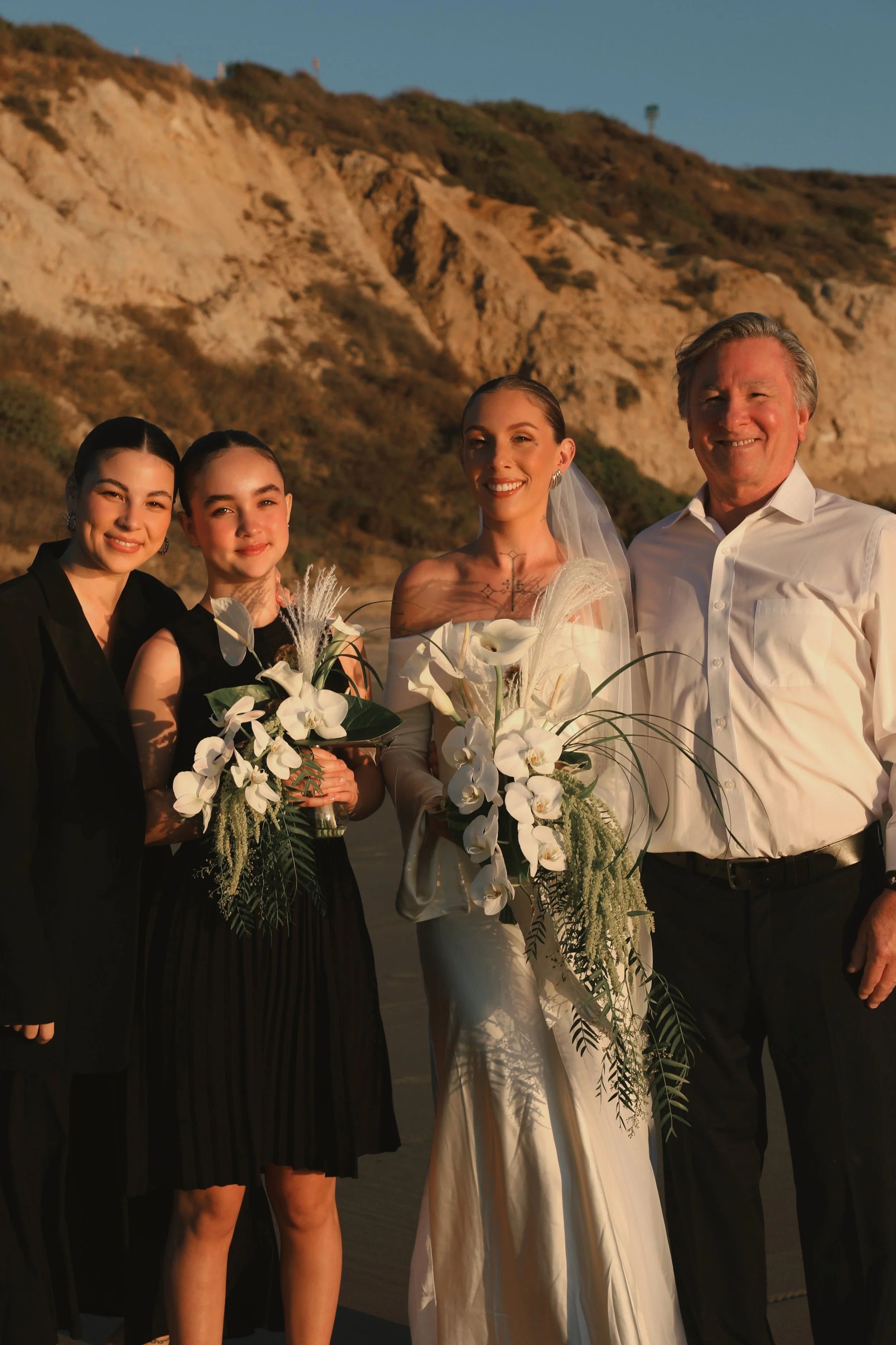 A group of four people, including a bride in a white wedding dress holding a bouquet and three women, stands outdoors against a hillside at sunset, smiling for a photo.