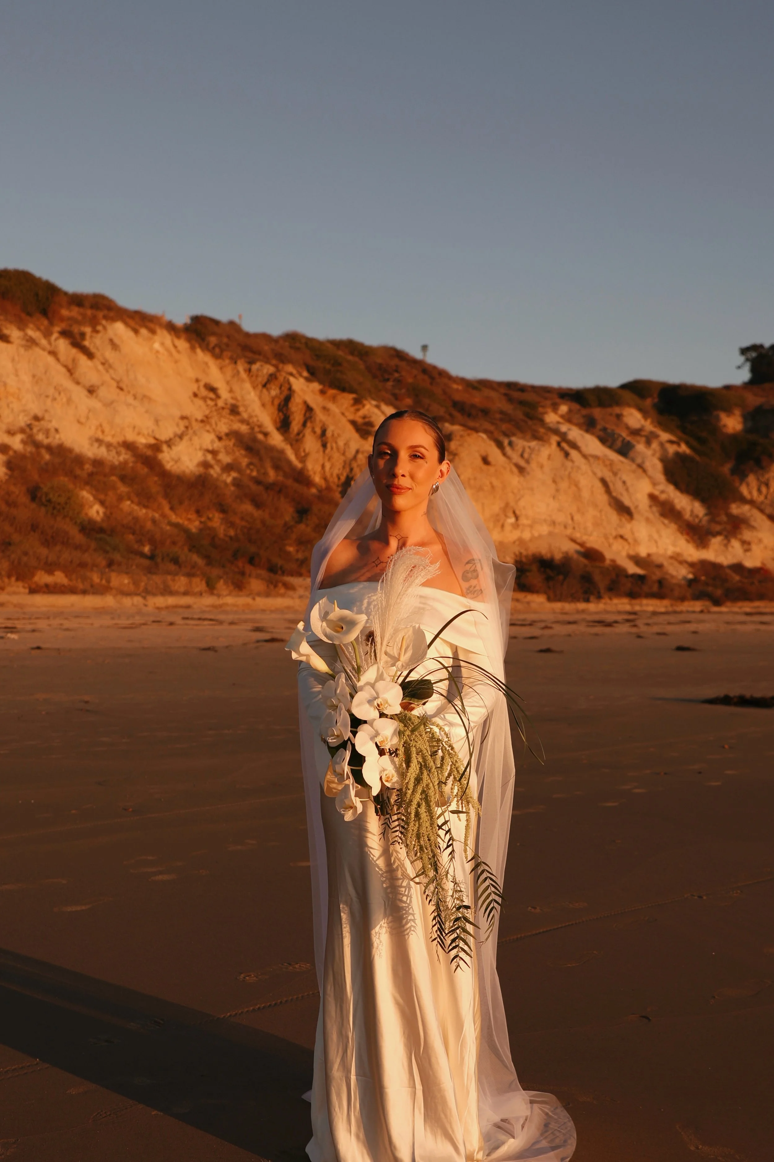 A woman in a white wedding dress holding a large bouquet of white flowers, standing on a beach during sunset with cliffs in the background.
