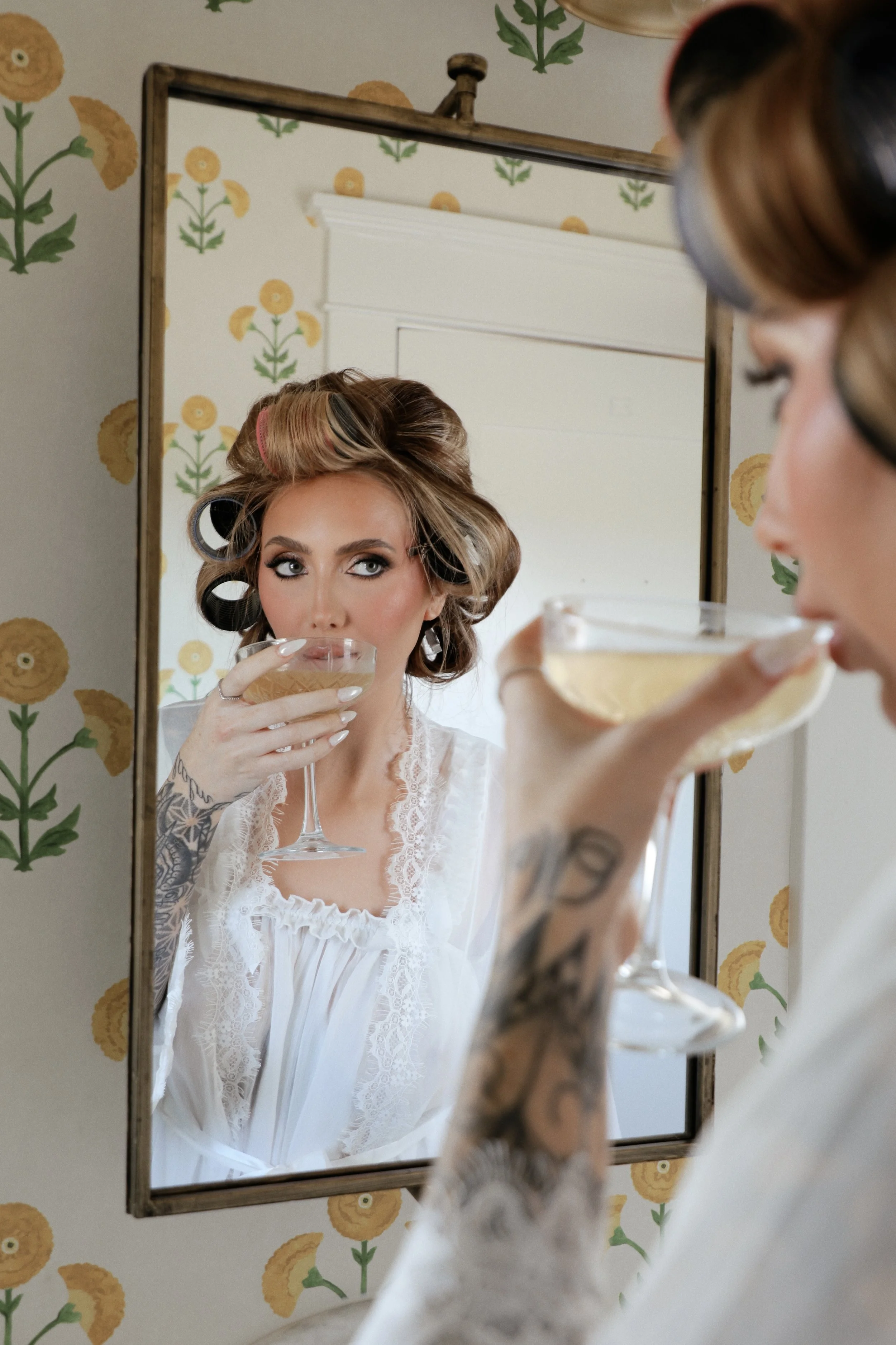A woman with curled hair and tattoos on her arm, dressed in a white lace robe, drinks champagne while looking at herself in a mirror hung on a wall with yellow and green floral wallpaper.