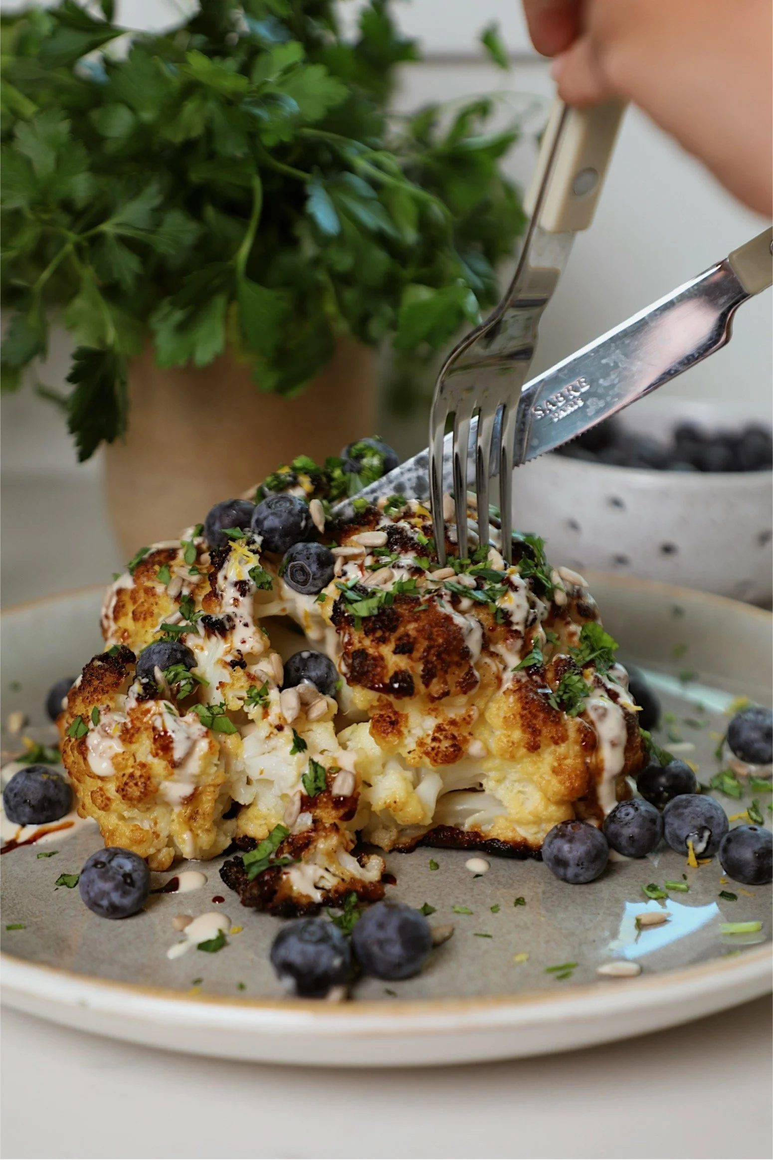 A plate of roasted cauliflower topped with blueberries, chopped herbs, and nuts, with a fork and knife placed in the cauliflower