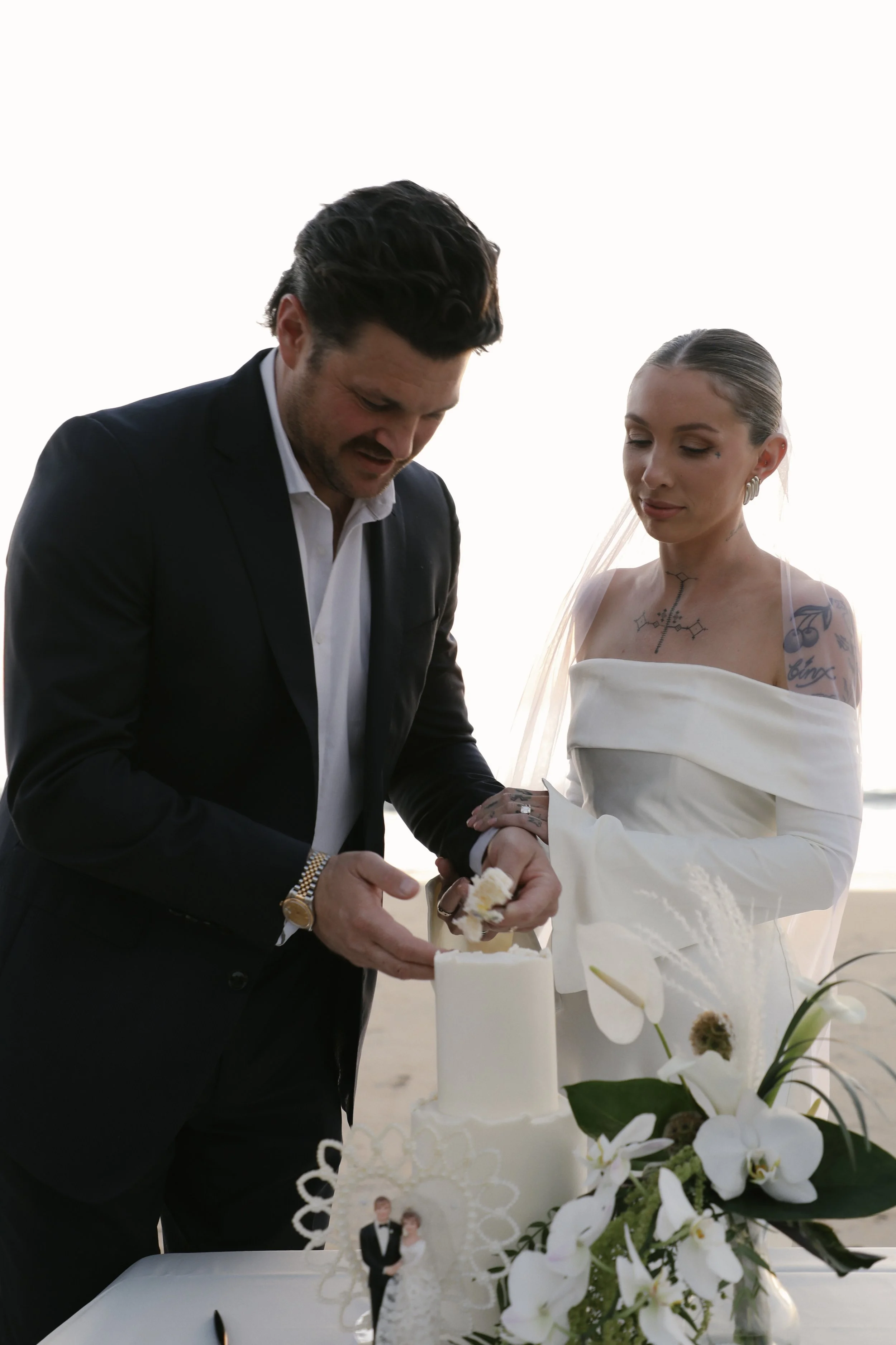 A groom and bride cutting a wedding cake on their wedding day, outdoors with a bright background.