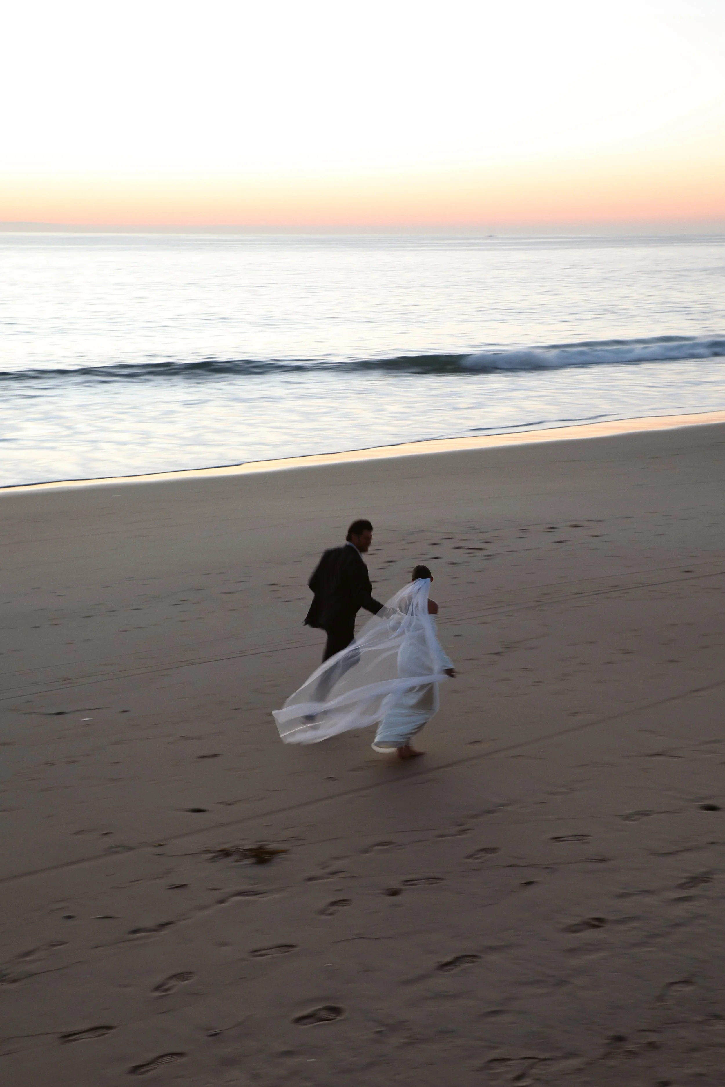 A couple in wedding attire walking on a sandy beach during sunset, with the ocean and colorful sky in the background.