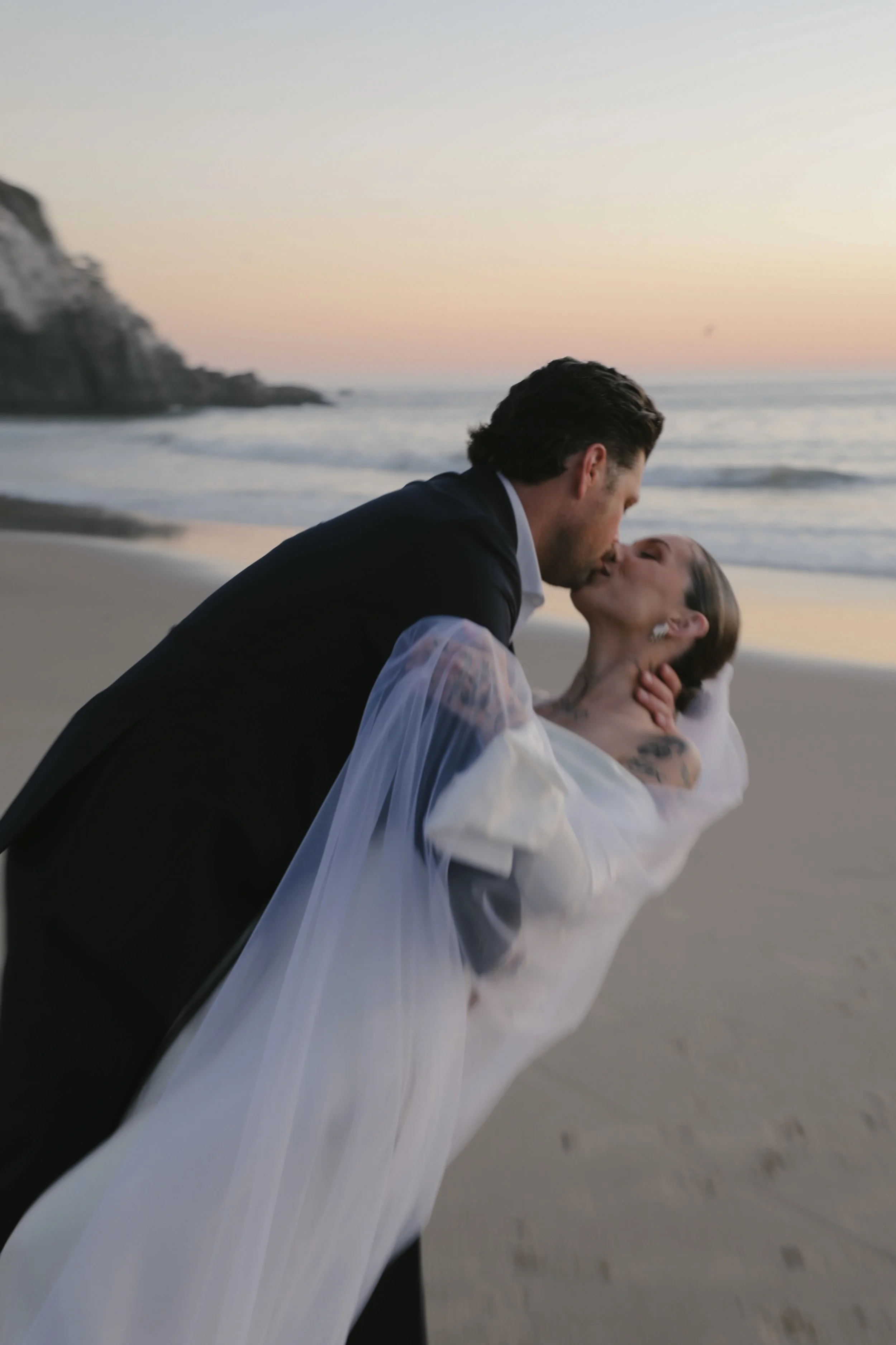 A bride and groom share a kiss on the beach at sunset, with cliffs visible in the background.