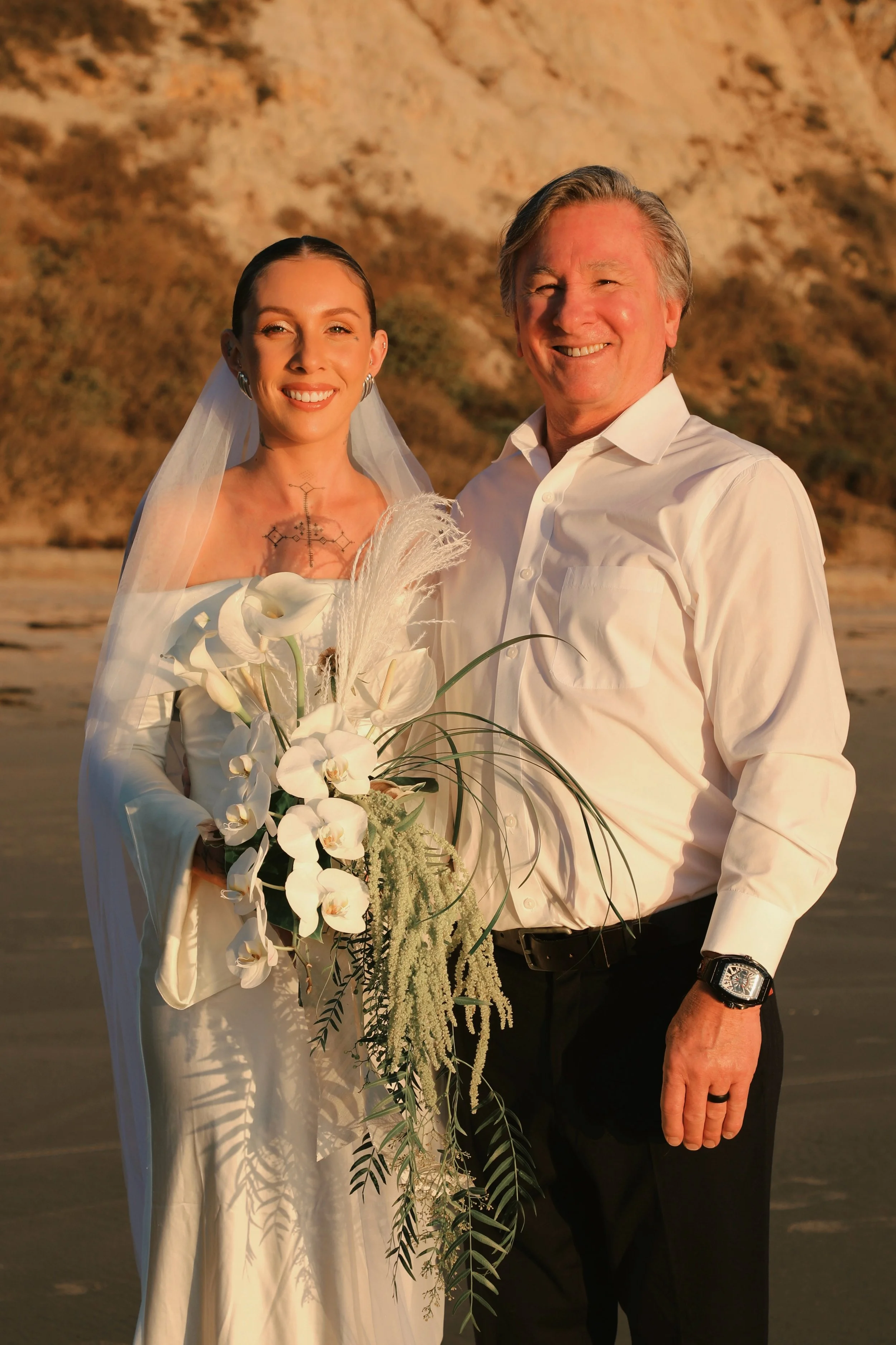 A bride and an older man standing on a beach at sunset, smiling for the camera. The bride is holding a large bouquet of white flowers and has a tattoo on her chest. She is dressed in a white wedding gown with a veil. The man is wearing a white dress