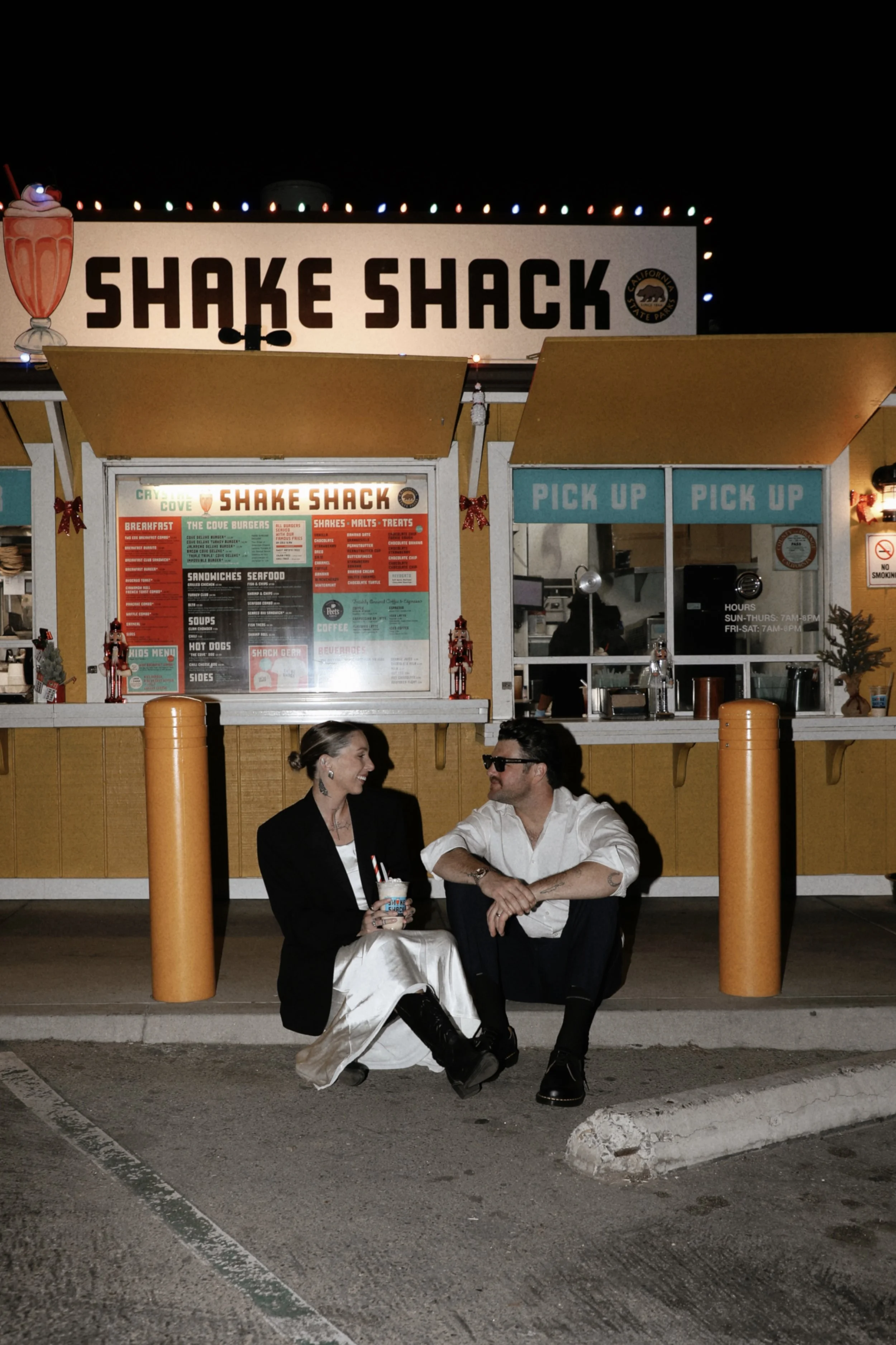 Two people sitting on the ground in front of Shake Shack, talking and smiling. The Shake Shack order window is visible behind them, with a menu and signs.