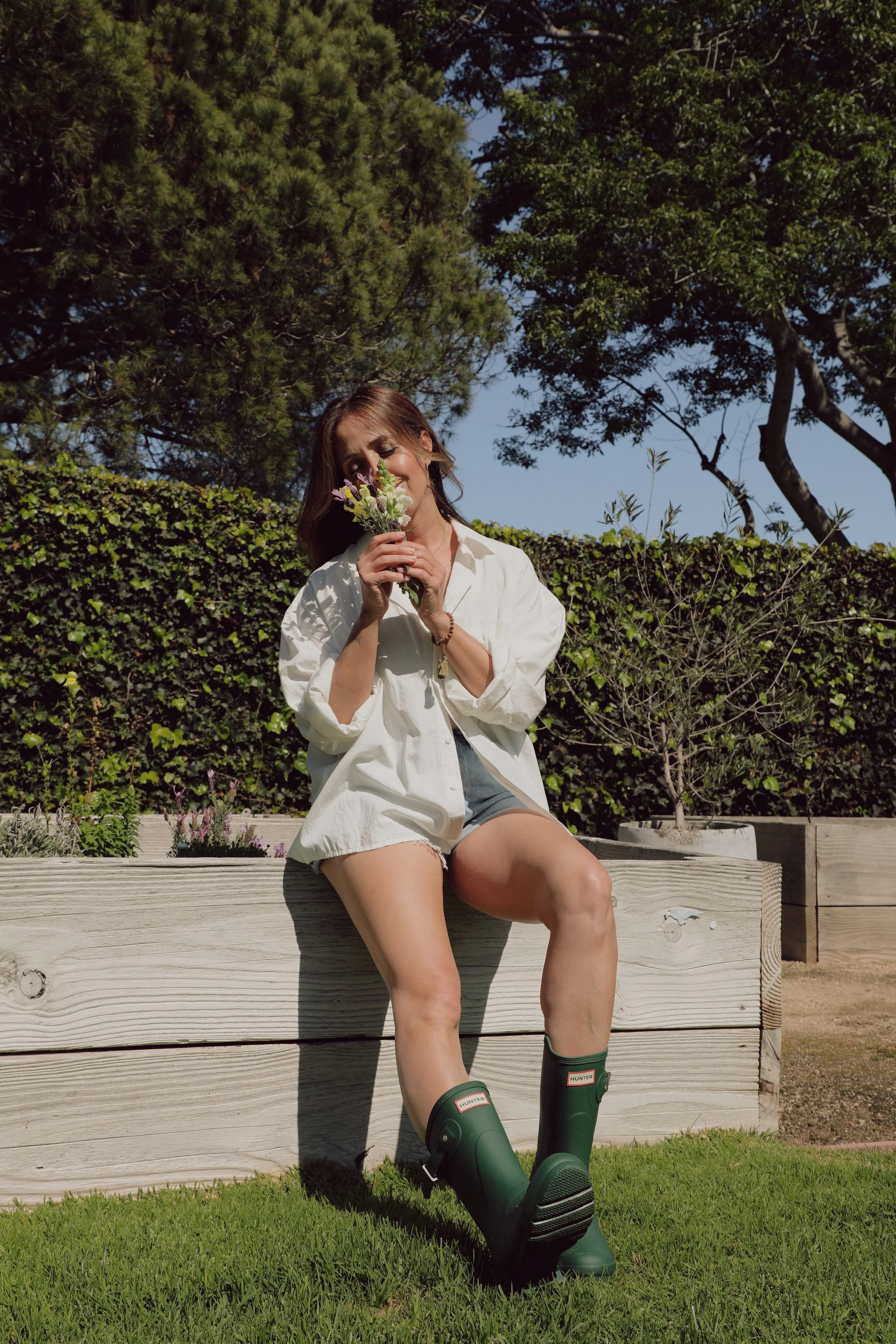 A young woman sitting on a wooden garden bed, wearing a white shirt, denim shorts, and green rain boots, holding a bouquet of flowers, outdoors with trees and bushes in the background.