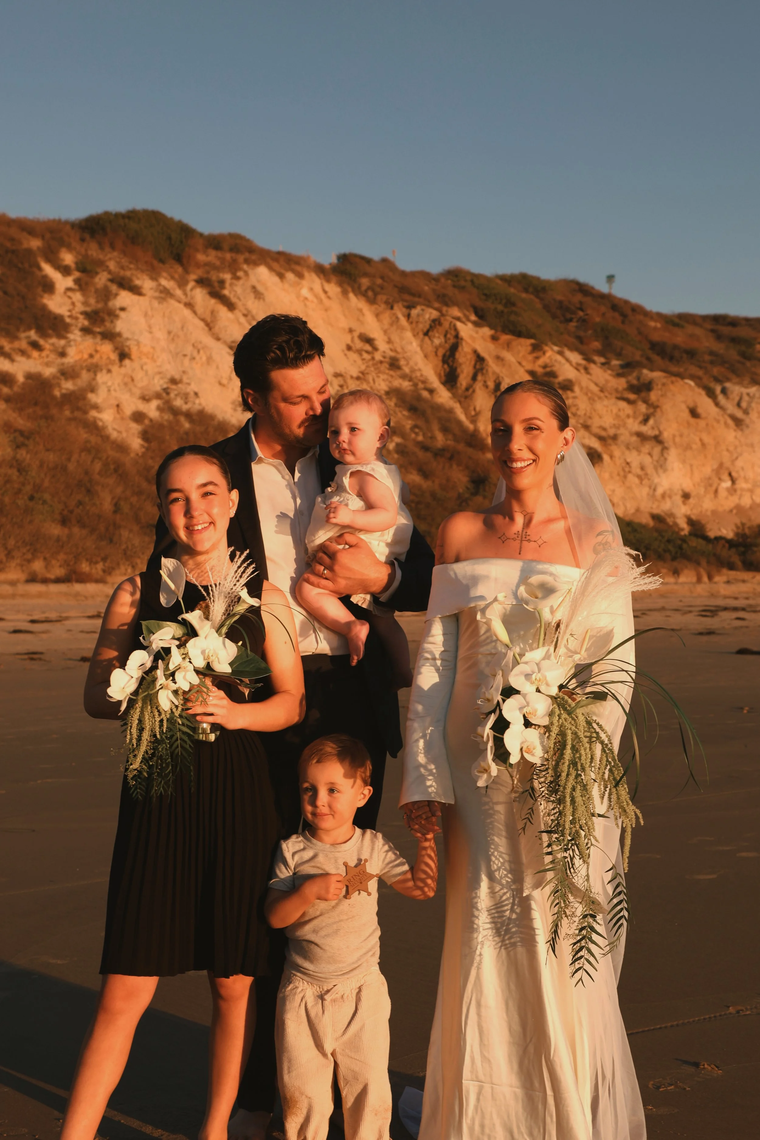 Family dressed in formal attire at a beach during sunset, holding flowers and a star-shaped object.