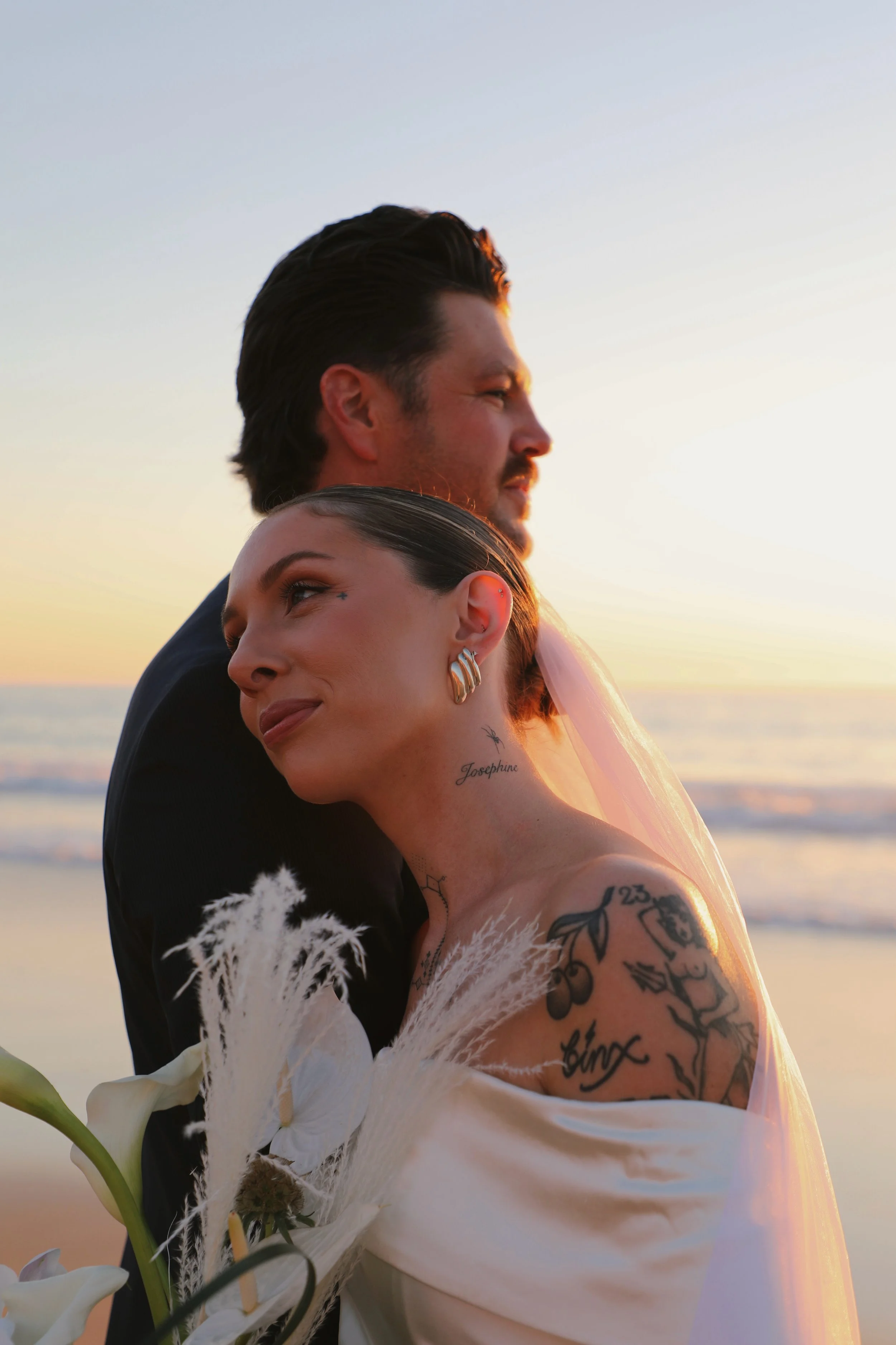 A bride and groom standing close together on the beach during sunset, with the bride smiling and holding a bouquet of white flowers and pampas grass, and the groom looking ahead.