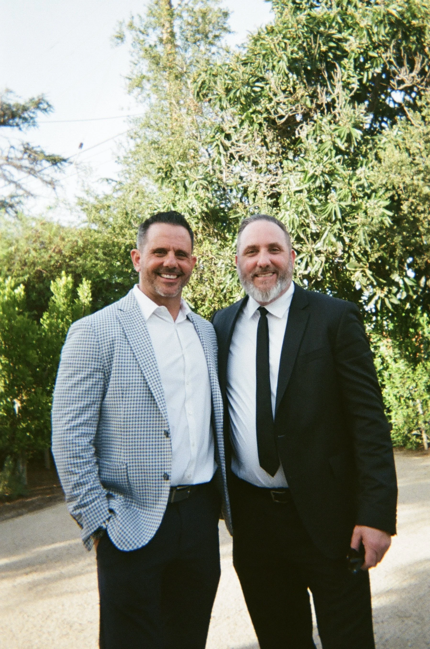 Two men in formal attire standing outdoors in front of trees, smiling at the camera.