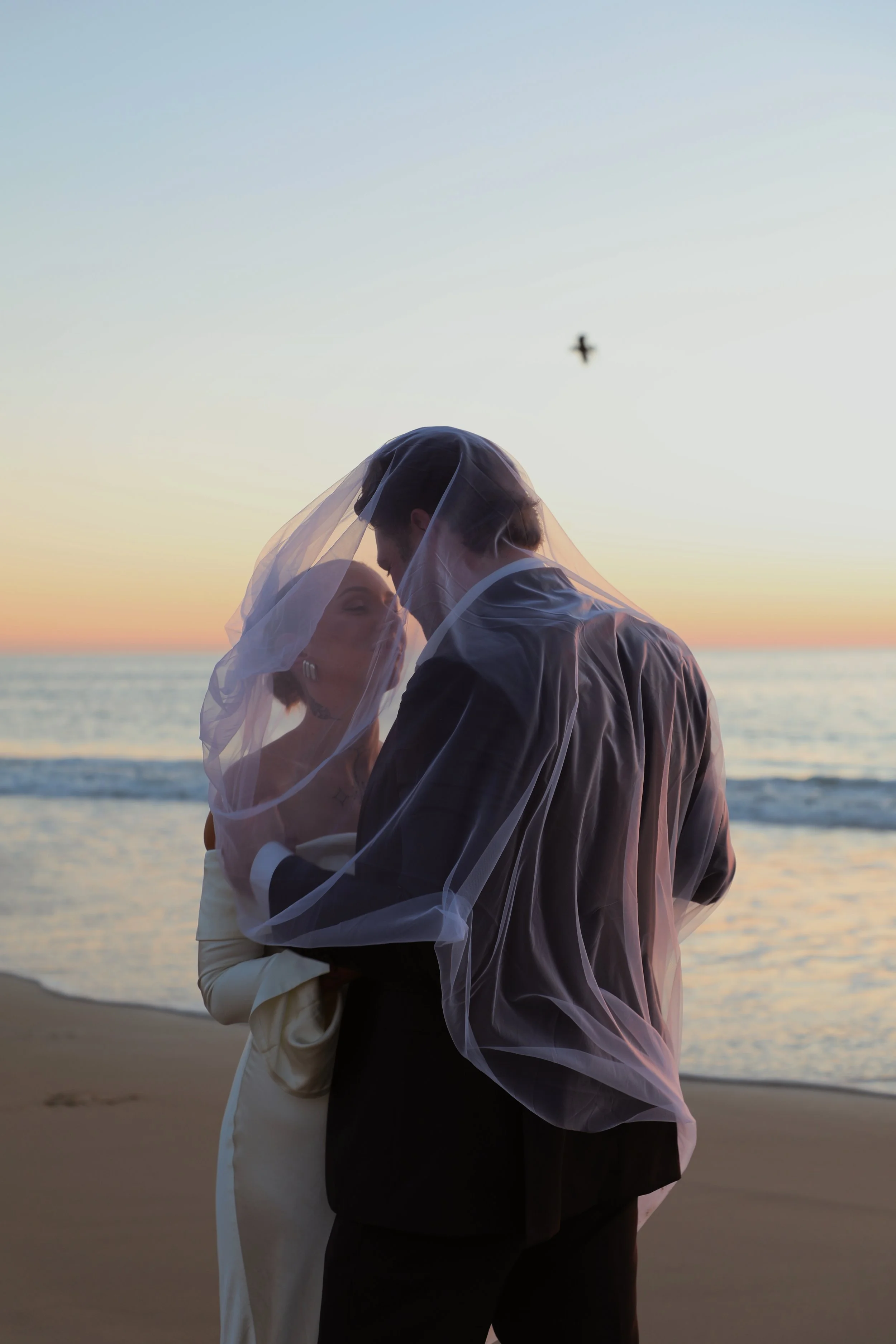 A couple dressed in wedding attire sharing a moment under a veil on the beach during sunset, with a bird flying in the sky.