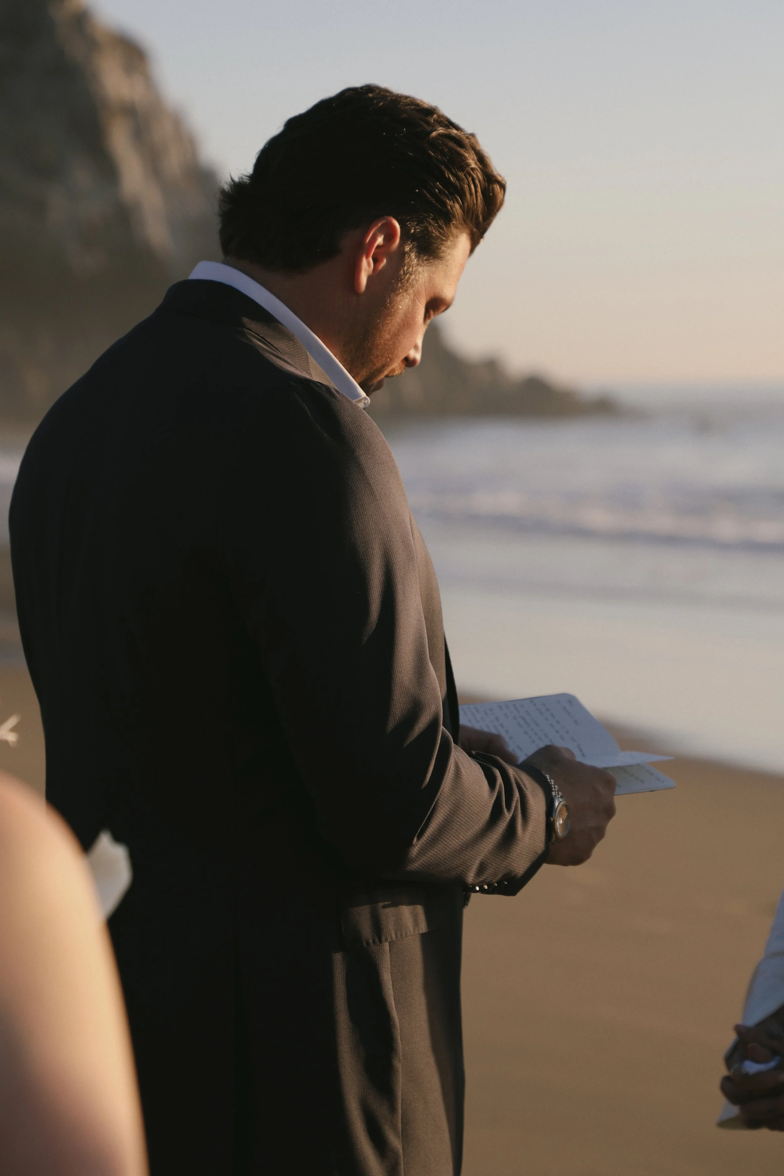 A man in a suit reading a book on a beach at sunset.