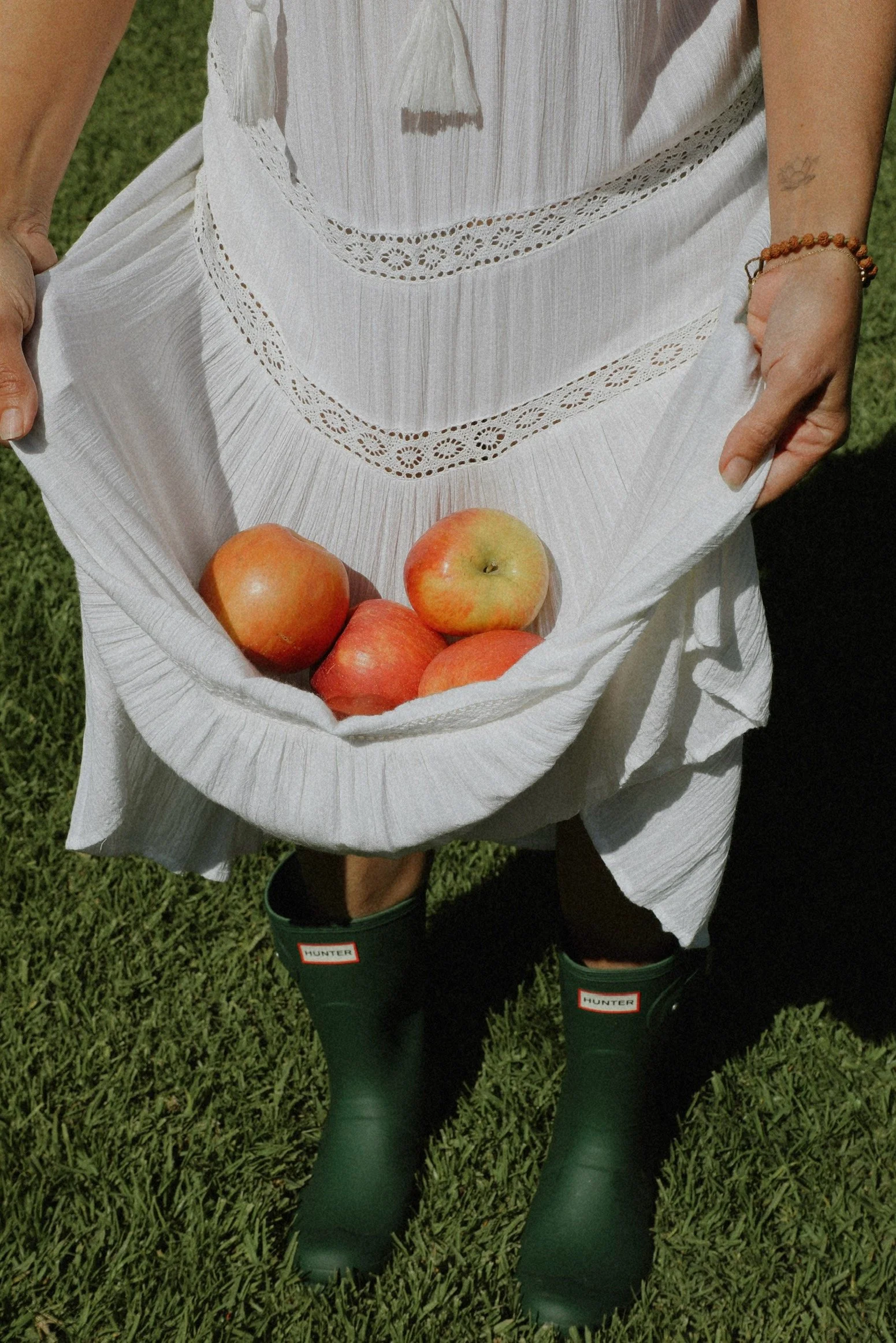 Person in a white dress holding a large cloth bag filled with red and yellow apples, standing on green grass, wearing green Hunter rain boots.