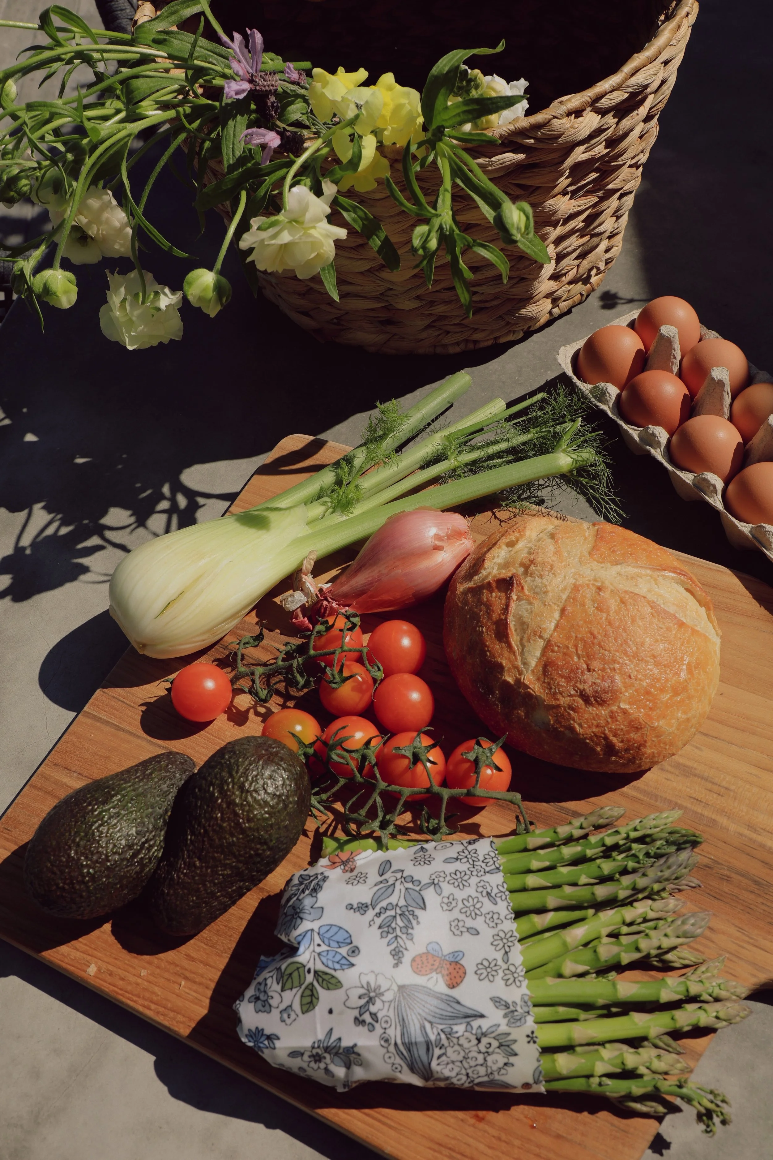 Fresh vegetables, eggs, and bread on a wooden cutting board with a basket of flowers nearby.