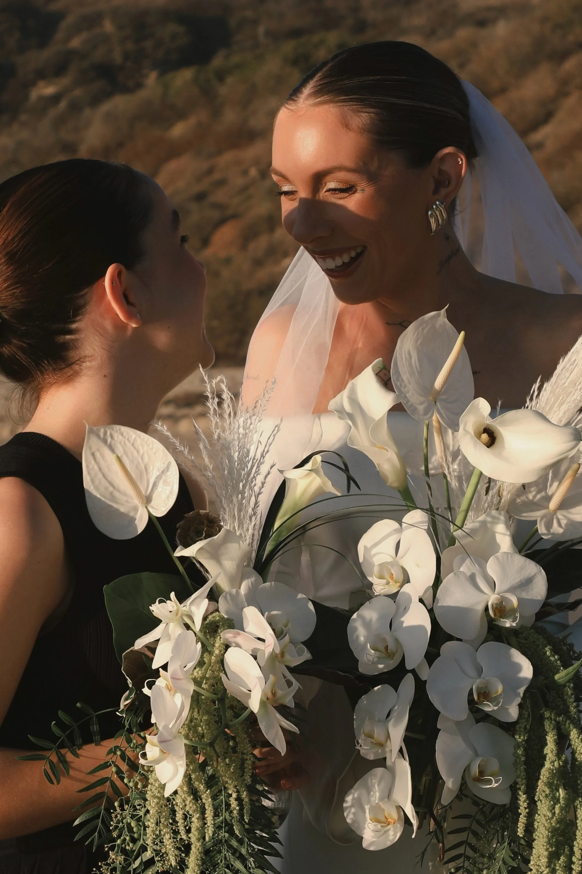 A bride in a wedding dress with a veil, holding a large bouquet of white orchids and greenery, smiling while looking at another woman in a black dress with her hair slicked back, outdoors with a blurred natural landscape background.