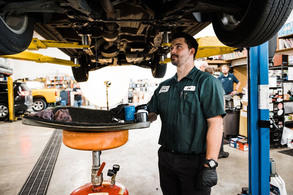 A man in a company’s shirt performing an auto repair, holding a car from underneath