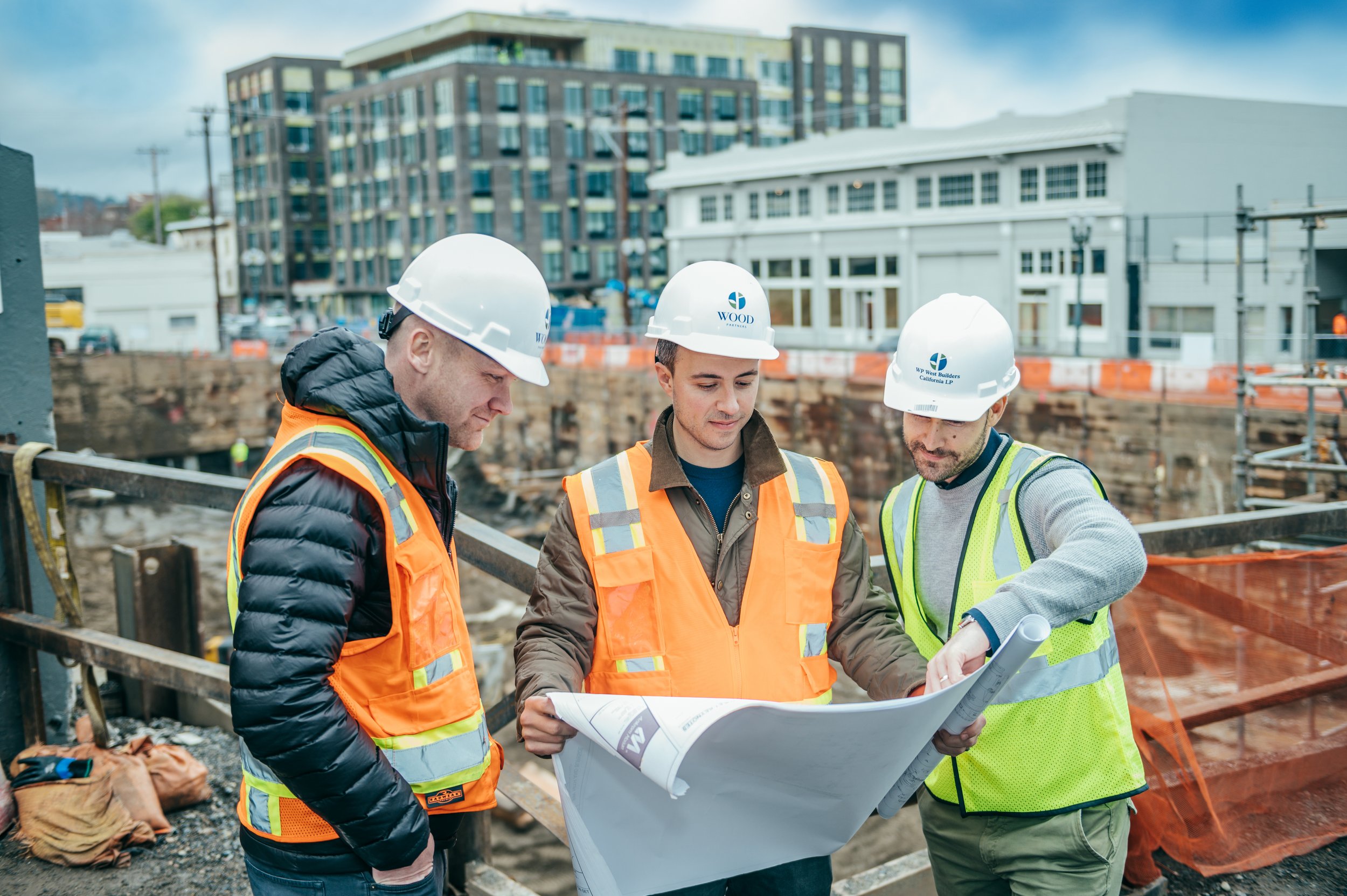 Three engineers in crash helmets and vests discussing plans at a construction site for property development.
